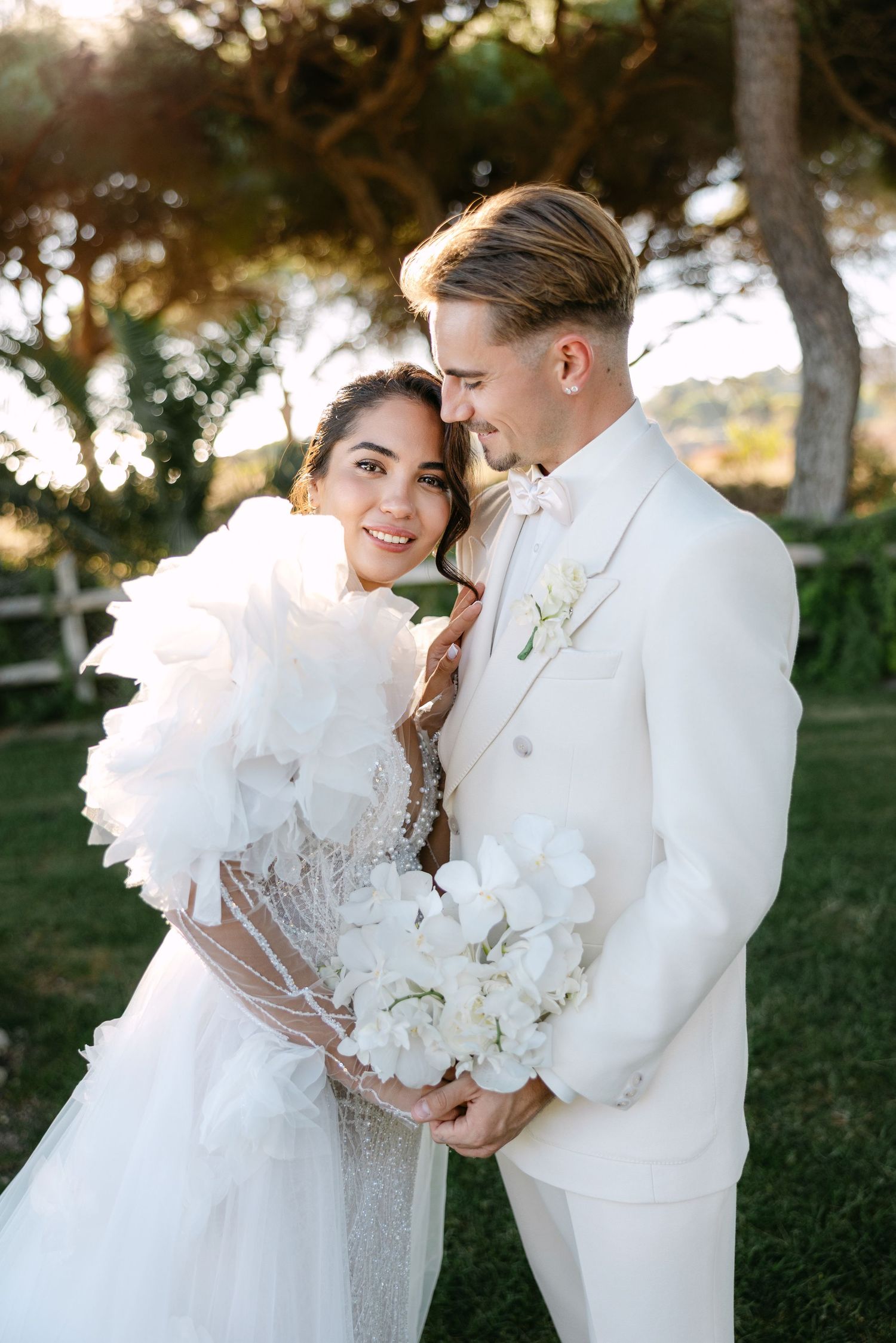 Romantic wedding couple in white attire embracing outdoors during golden hour.