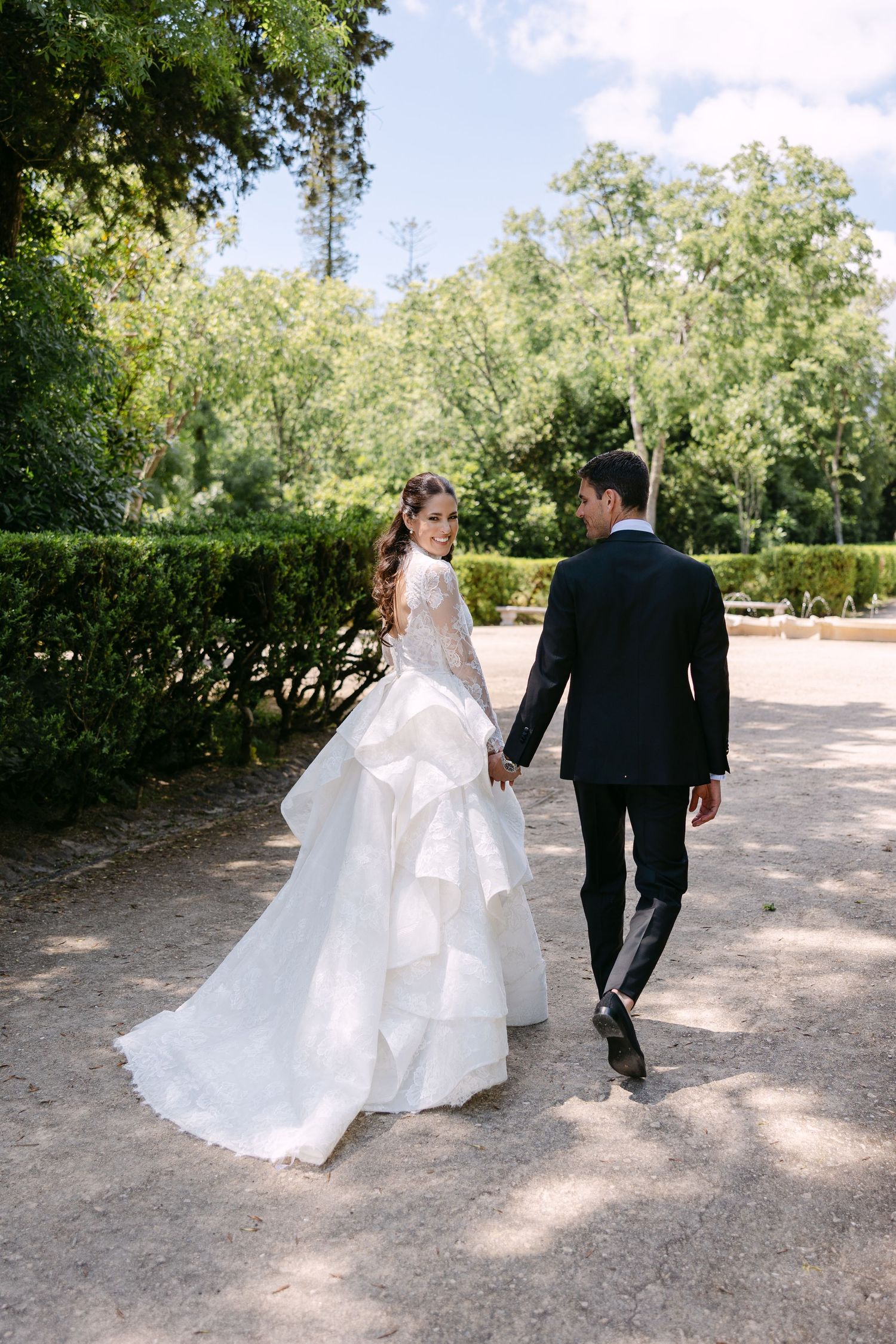 Bride in white gown and groom in black tuxedo walk hand in hand through a lush garden path.