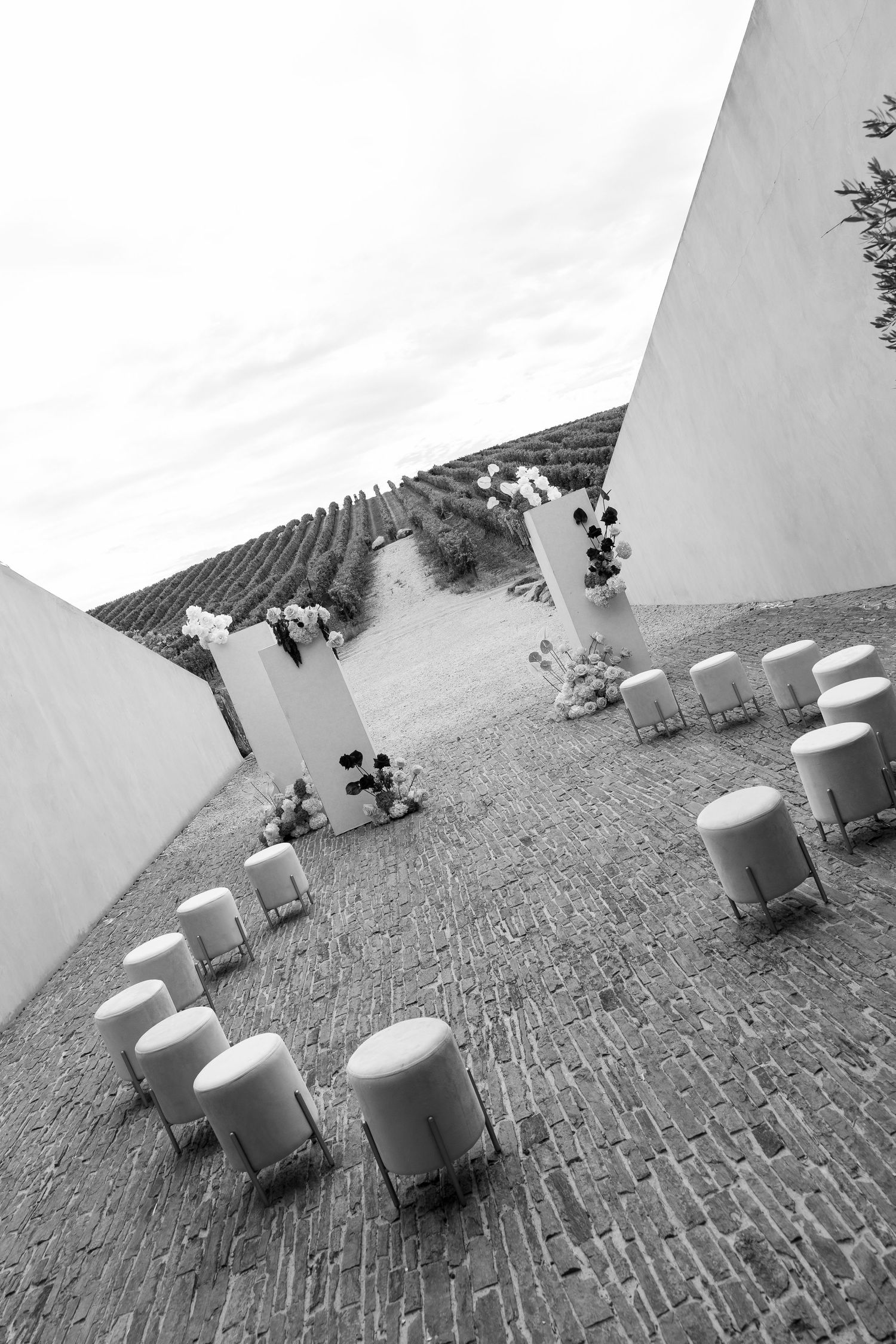Black and white aerial view of an outdoor wedding ceremony setup with round stools on cobblestone.