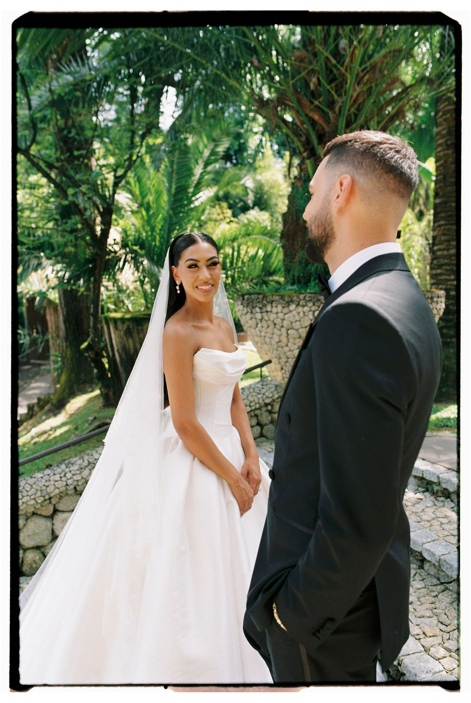 Bride in strapless white gown with veil faces groom in black tuxedo outdoors amid lush tropical greenery.