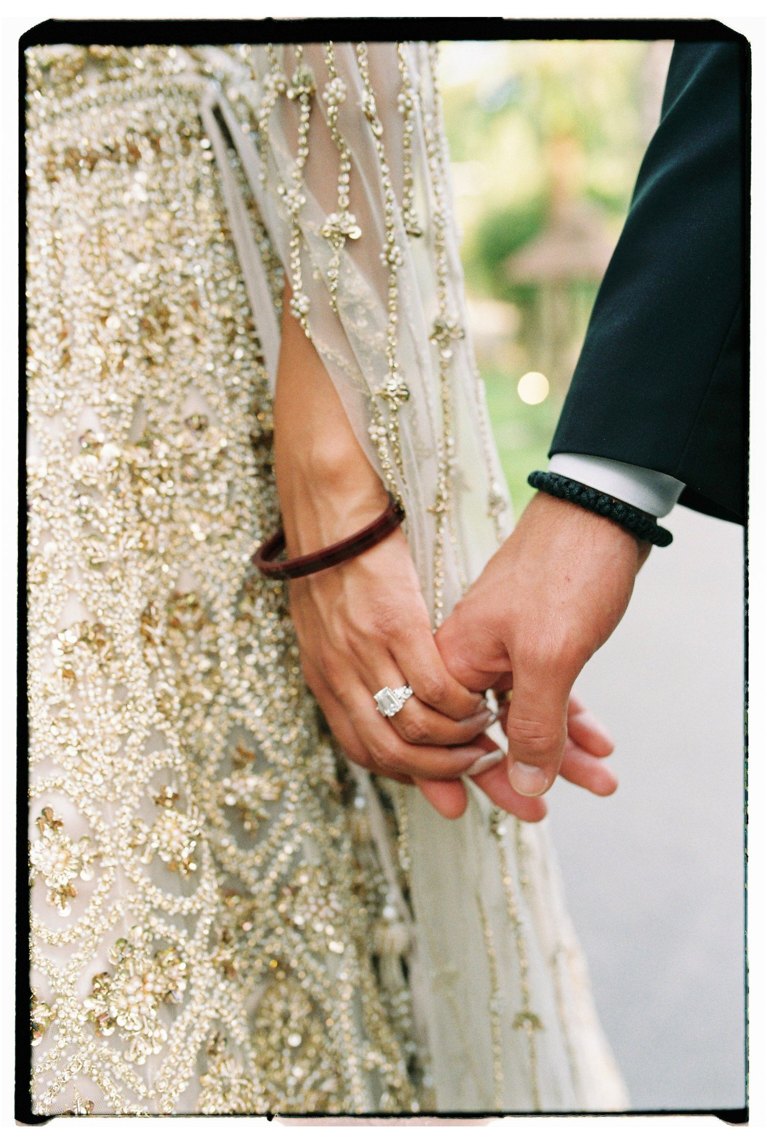 Couple holding hands, woman wearing diamond ring and gold embroidered bridal outfit, man in navy suit with bracelet.
