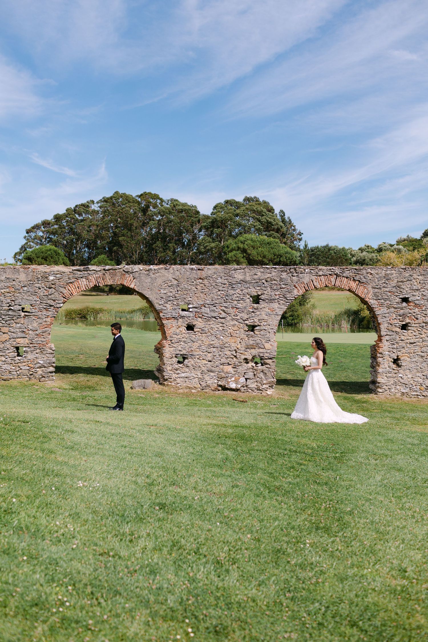 Wedding couple poses playfully in front of historic stone archways in a grassy field under blue skies.