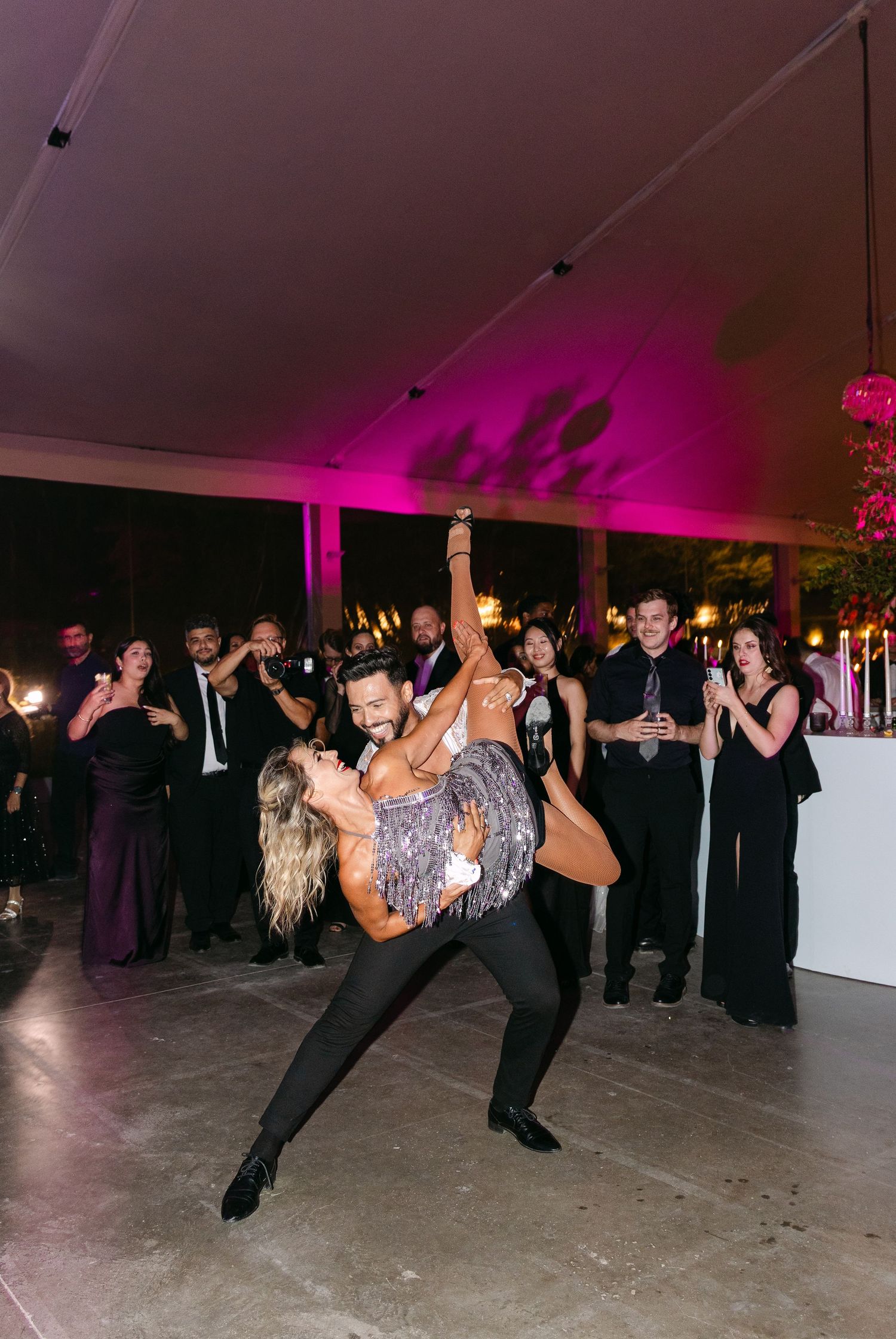 Wedding reception dance floor filled with guests performing synchronized dance moves under pink lighting.