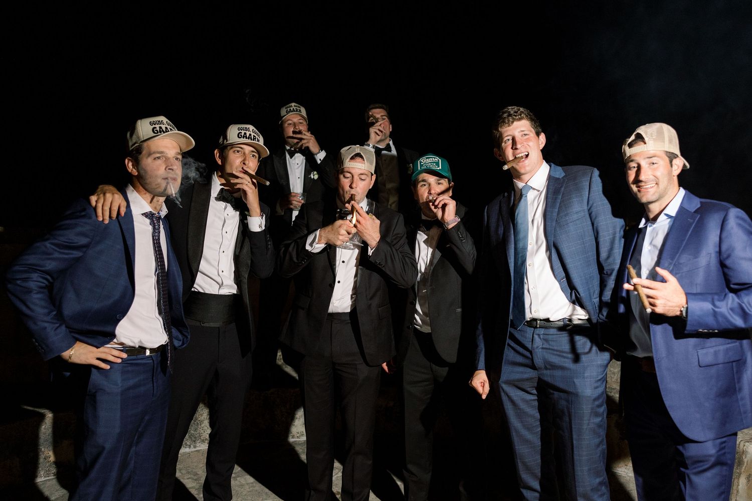 Group of groomsmen in suits wearing baseball caps posing together at night.
