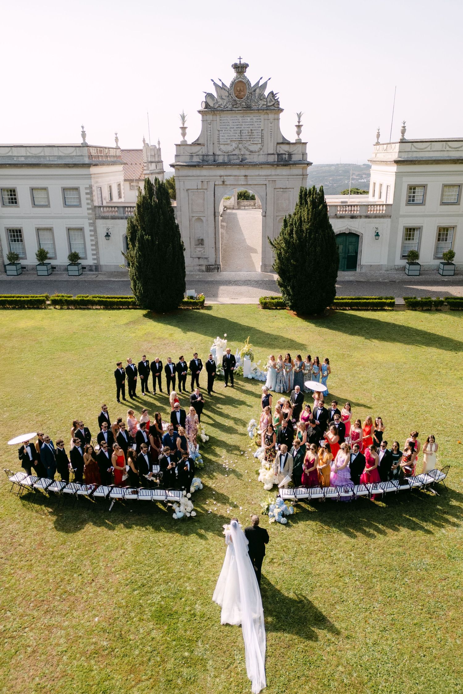Aerial view of wedding guests arranged in heart shape formation on lawn in front of ornate white building.