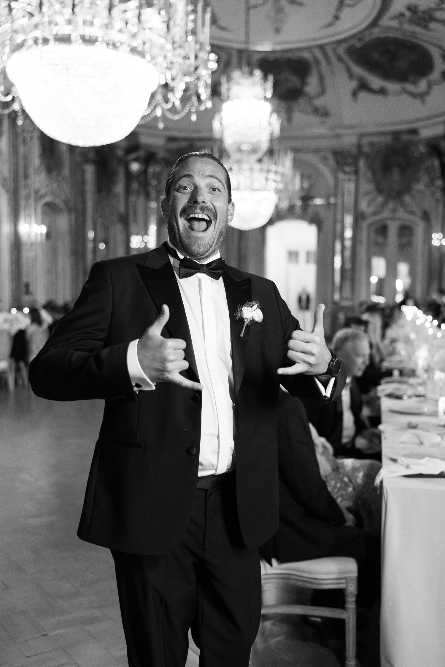 Joyful man in tuxedo giving thumbs up in an ornate ballroom with crystal chandeliers.