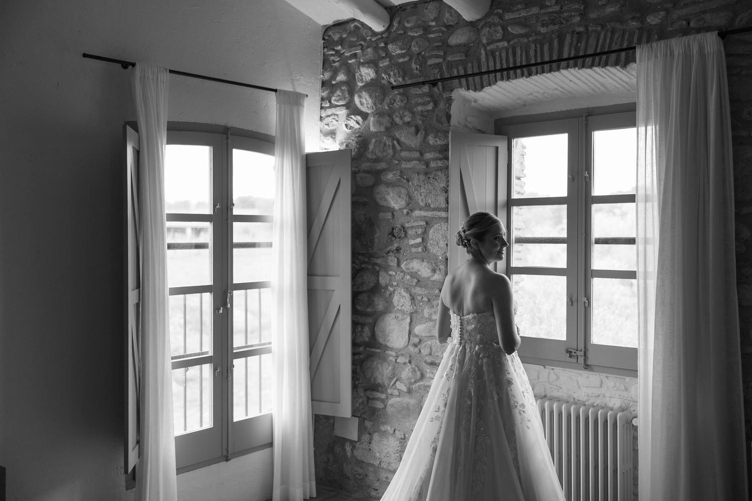 Black and white photo of a wedding dress hanging in a rustic stone room with natural light.