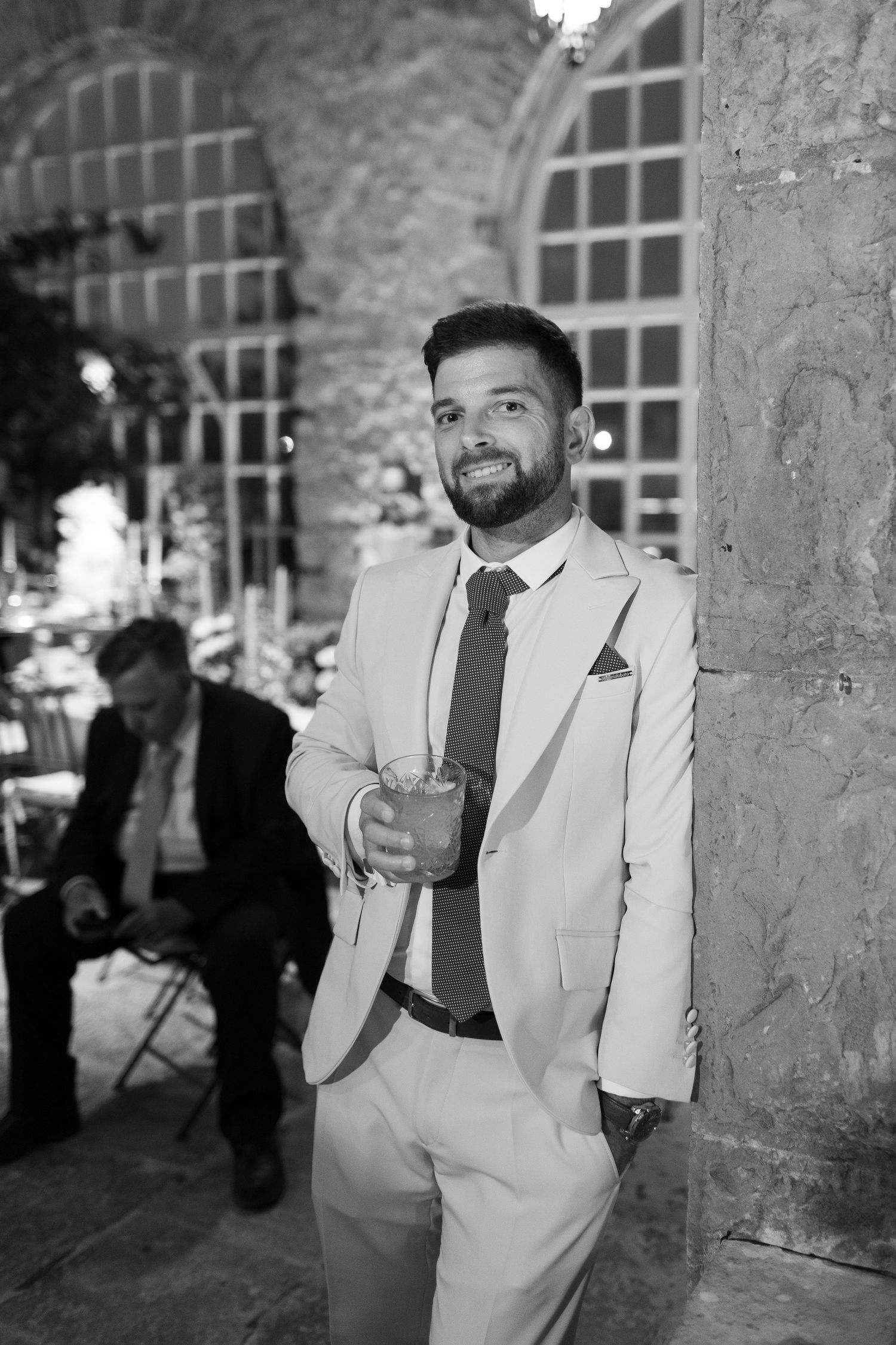 A guest in a light colored suit poses against a stone wall with arched windows at a wedding venue.