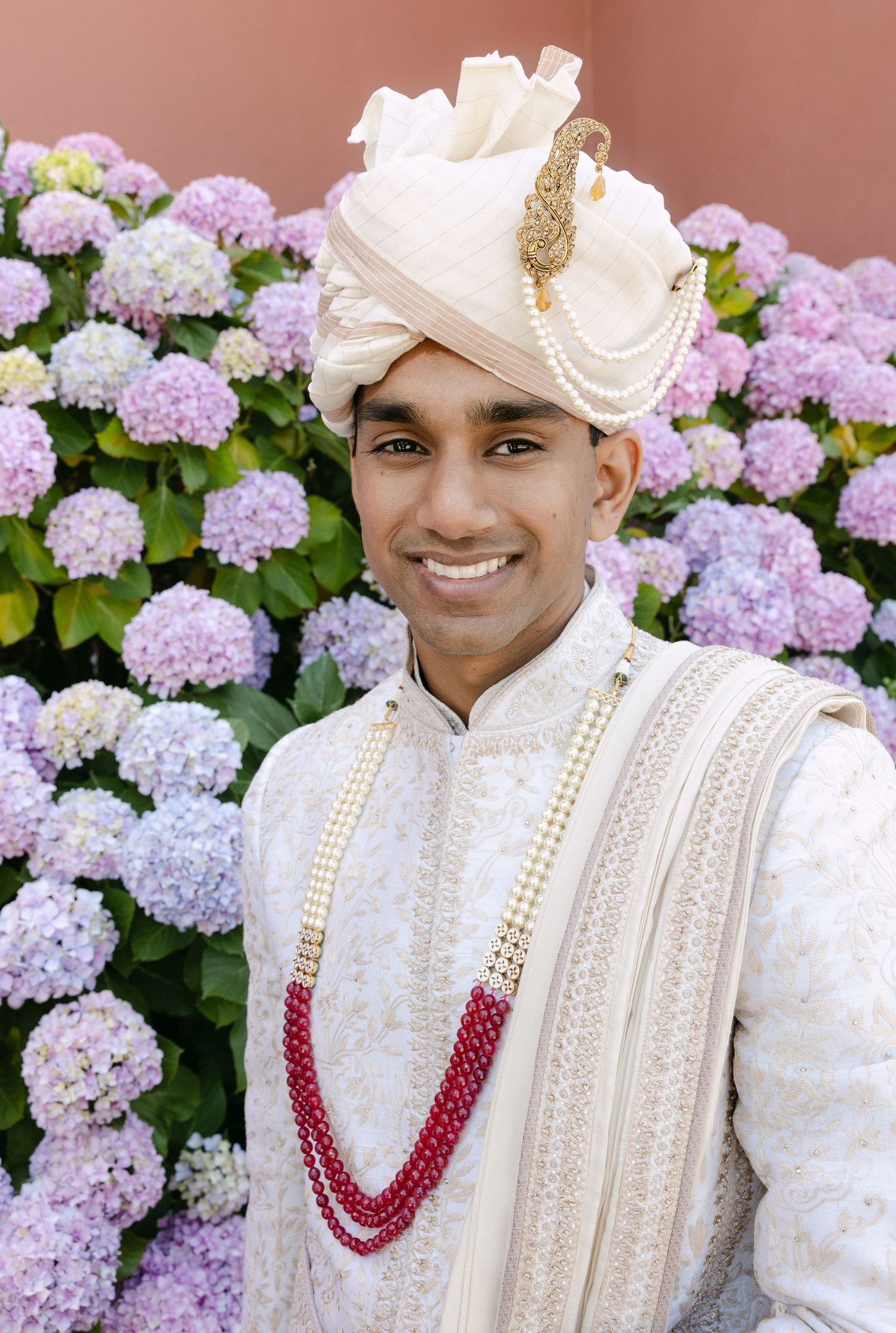 Traditional Indian groom wearing white sherwani and red beaded necklace poses against purple hydrangea flowers.