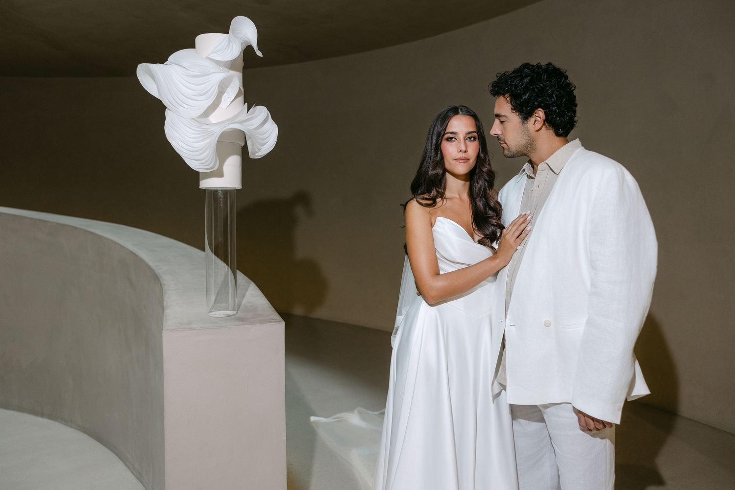 Couple in white attire standing beside abstract white sculpture in modern curved interior space.