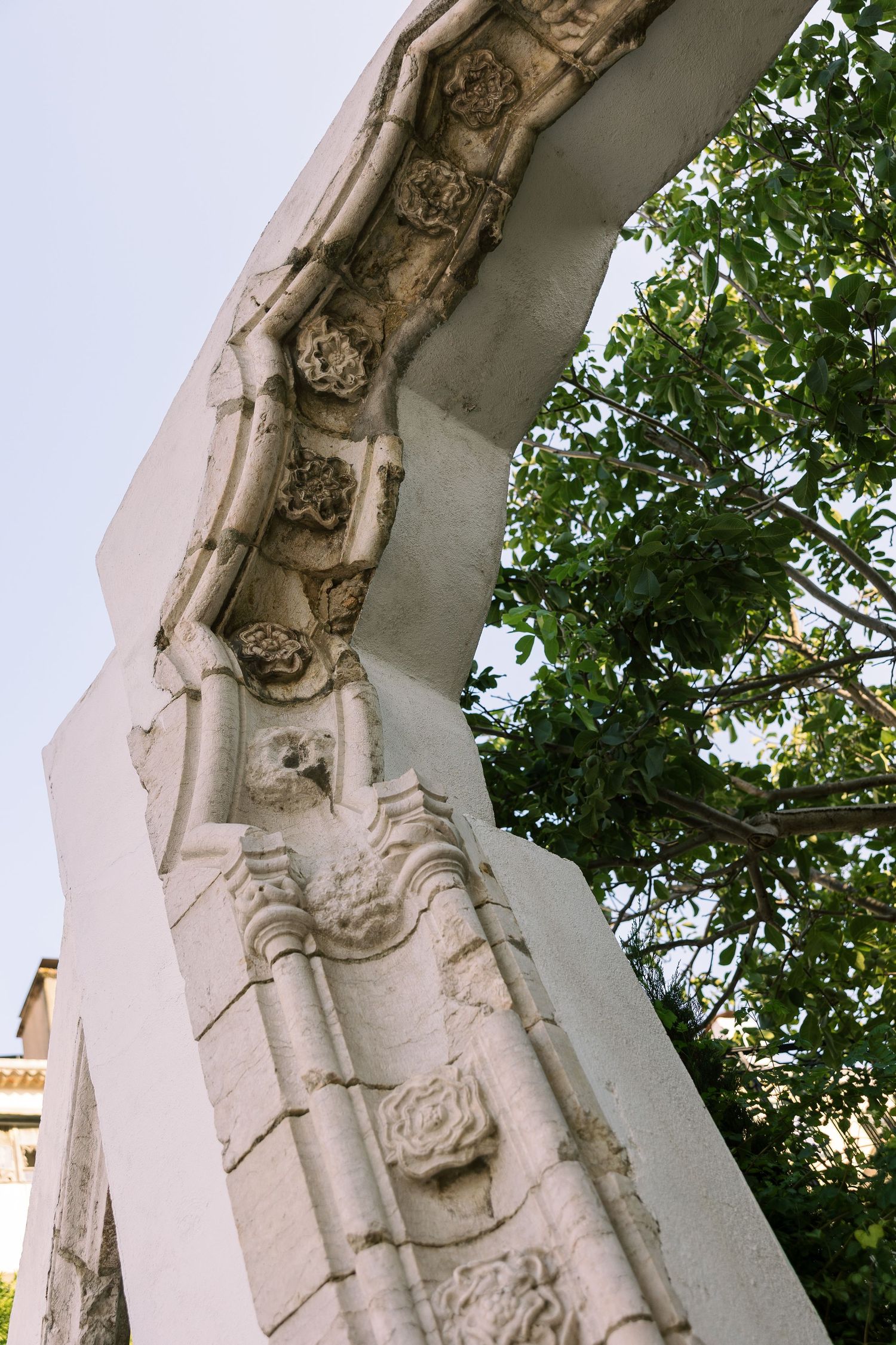 Ornate white stone arch with intricate carved floral details and green tree foliage against a clear sky.