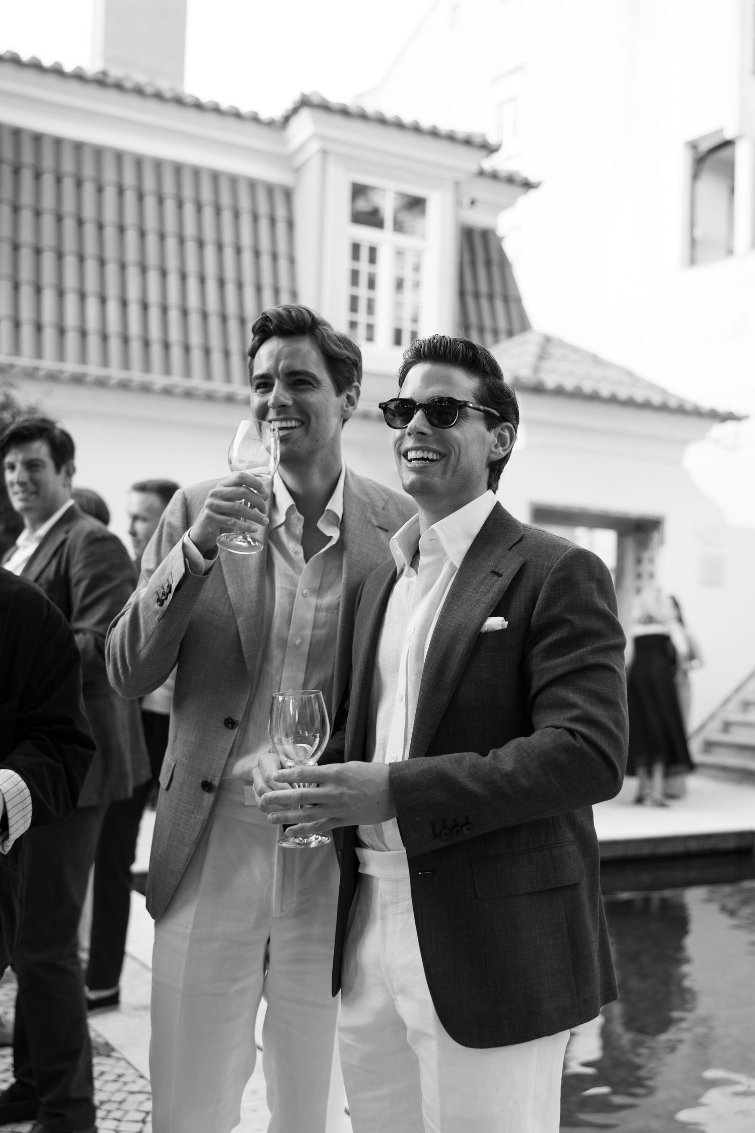 Two well-dressed men in blazers laughing and holding wine glasses at an outdoor social event.