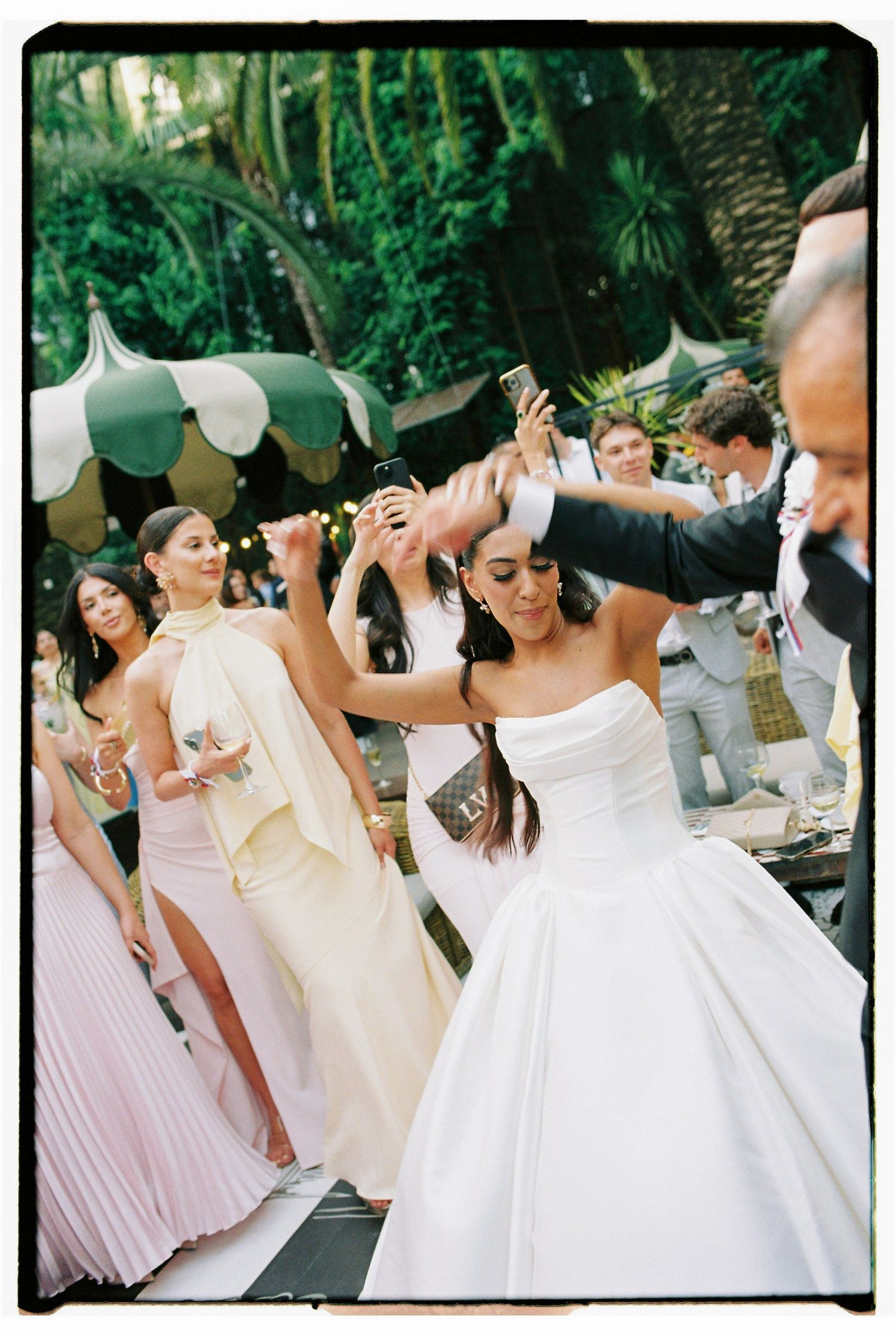 Bride in strapless white ball gown dances joyfully with guests at an outdoor garden wedding reception.