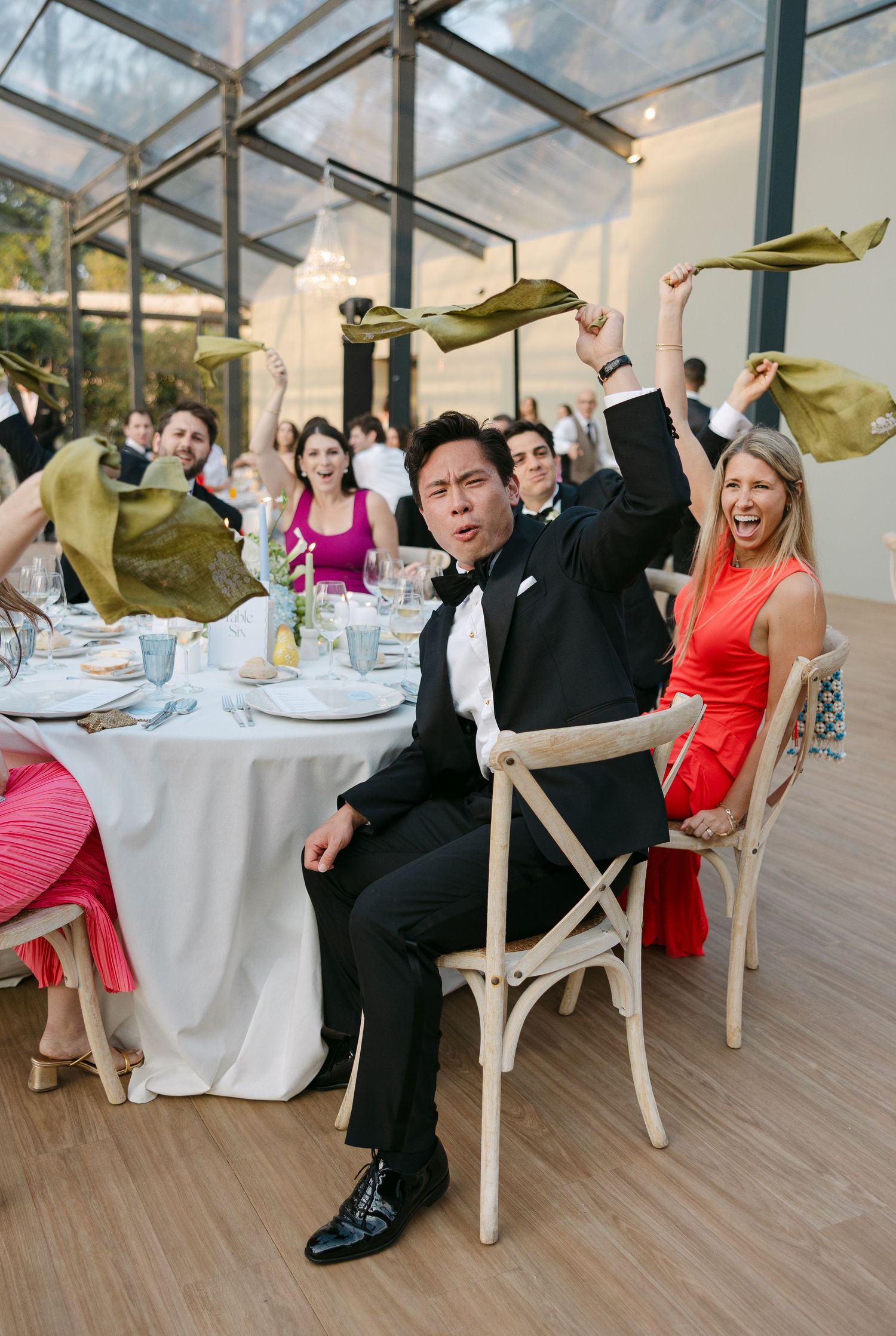 Wedding guests in formal attire celebrating enthusiastically at a glass-enclosed reception venue.