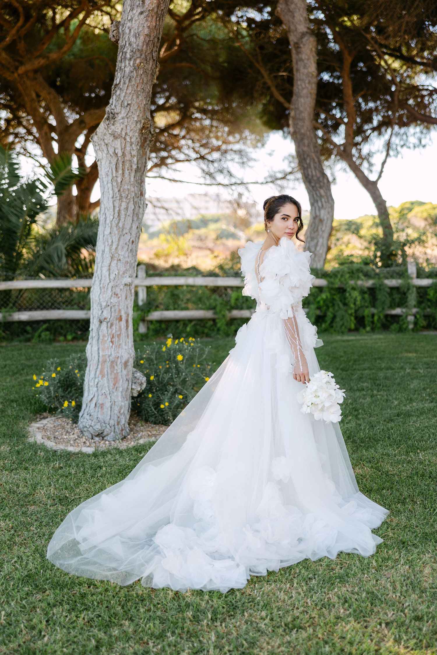 Flowing white wedding gown displayed in a garden setting with pine trees.
