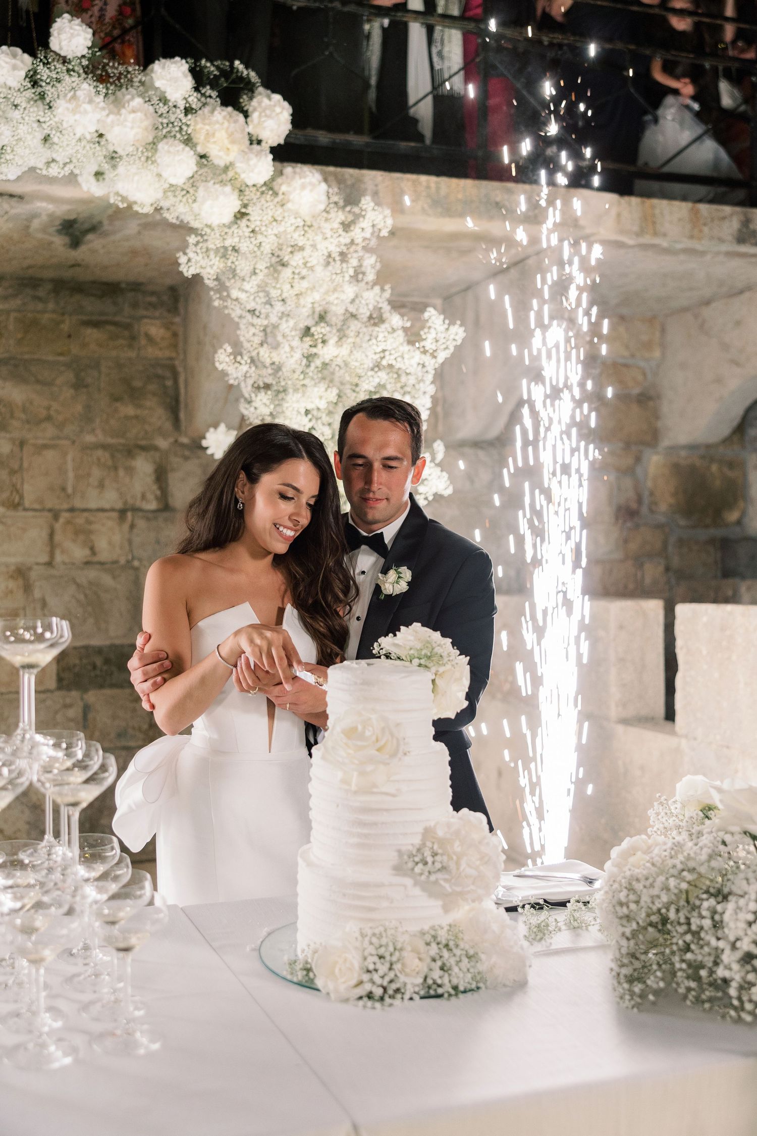 Wedding cake cutting moment with sparklers and white floral decorations in stone venue.