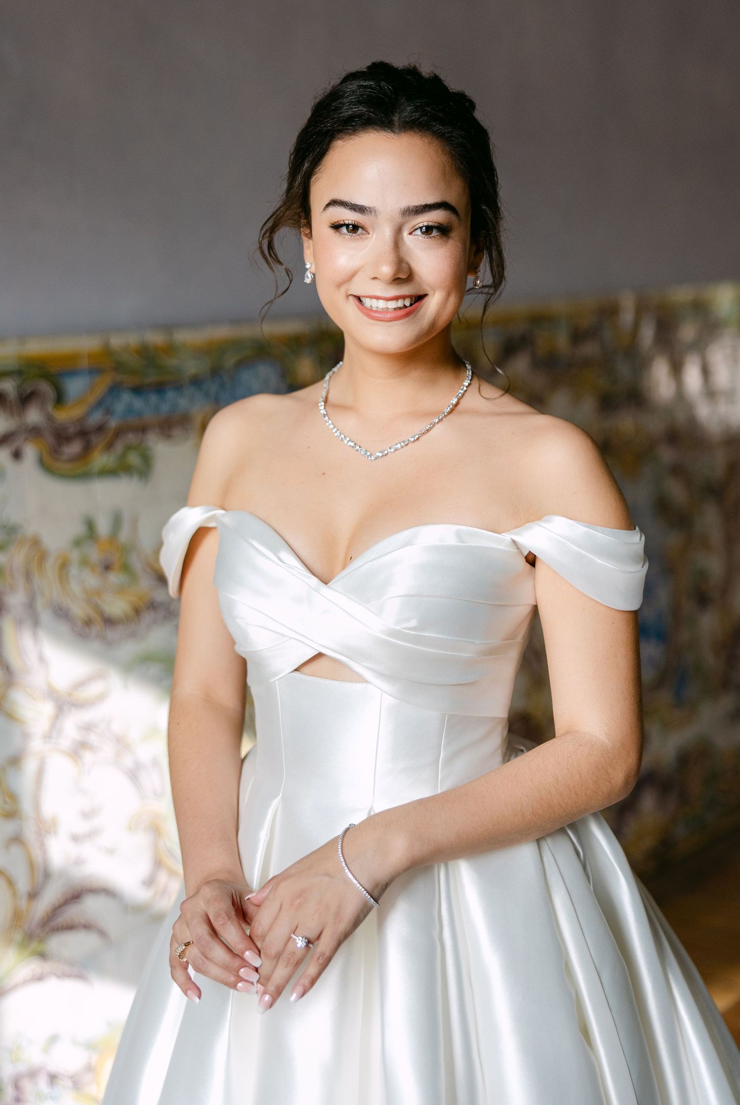 A bride in a classic white off-shoulder wedding gown stands gracefully against a decorative background.