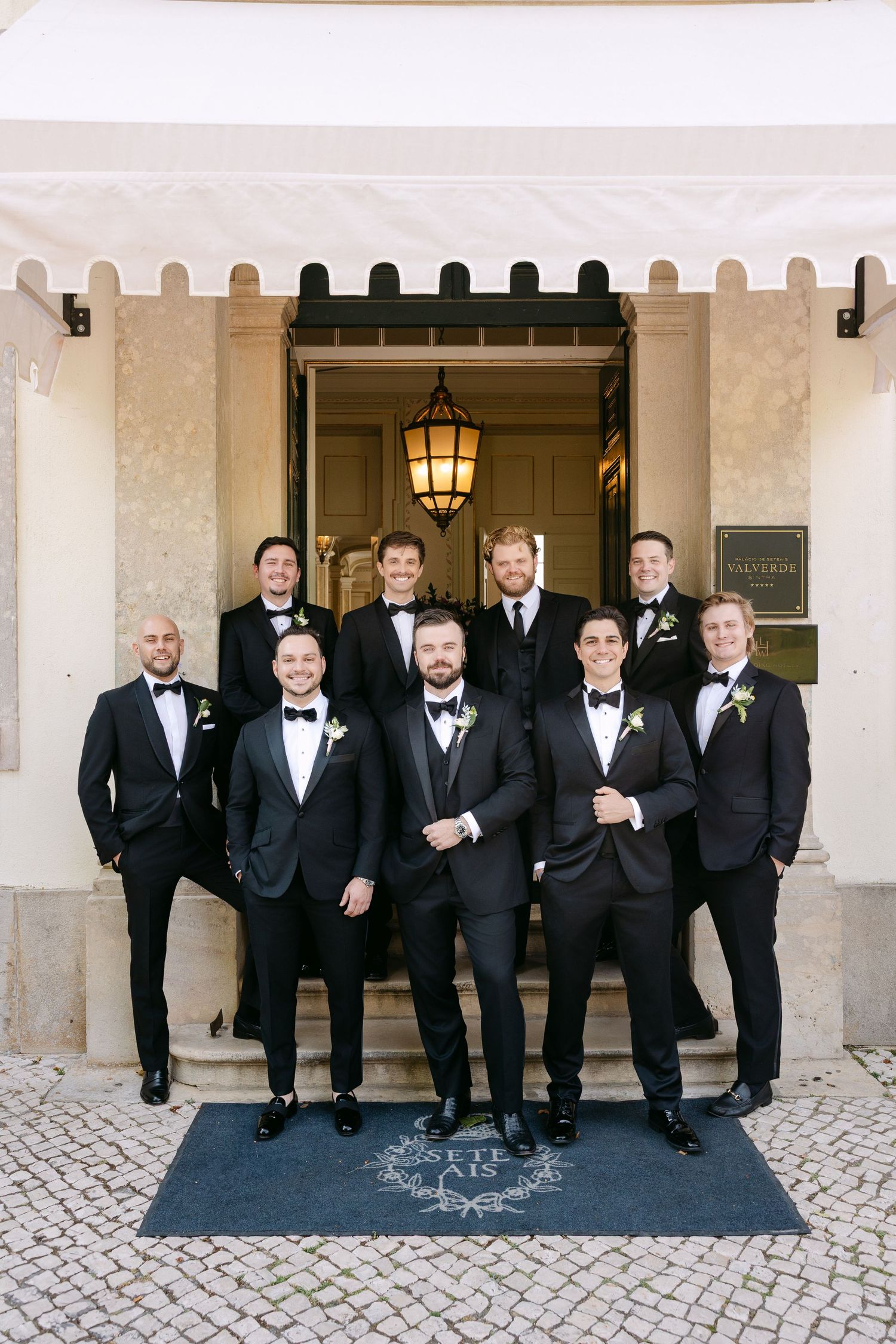 Groomsmen in black tuxedos pose together on a stone patio for formal wedding photos.