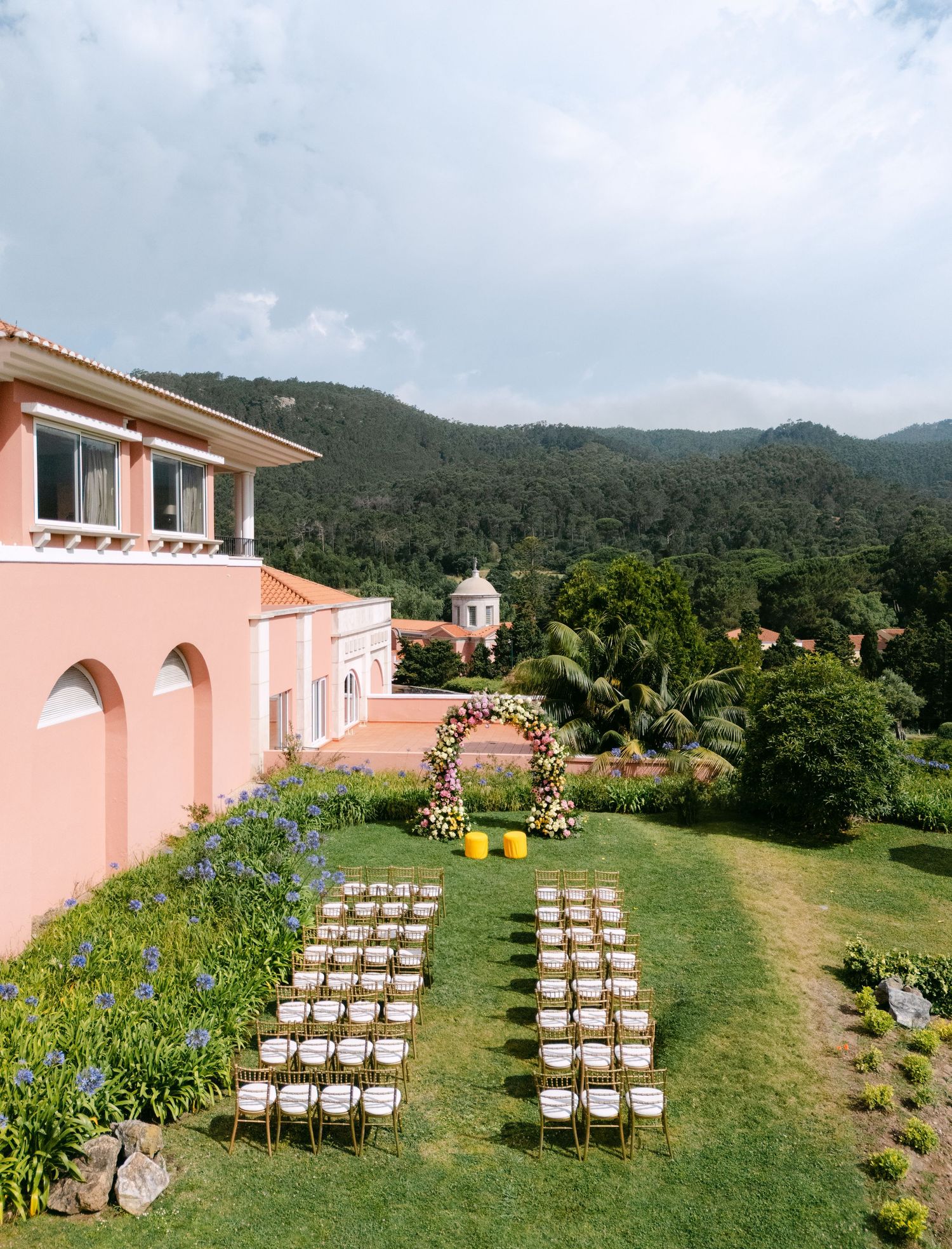Wedding ceremony setup with white chairs on a lawn in front of a pink Mediterranean-style villa.