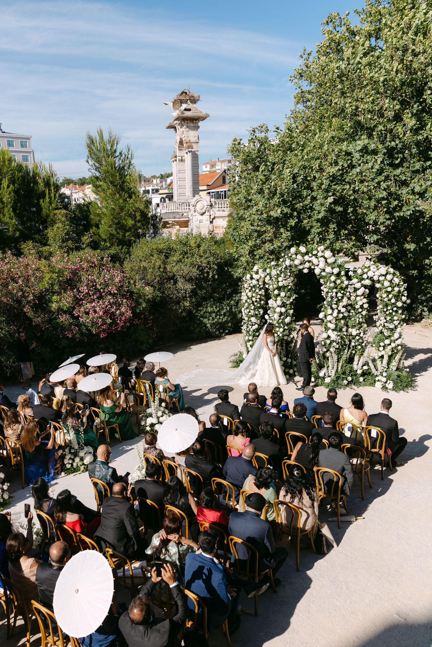 Wedding guests seated in wooden chairs during an outdoor ceremony with floral decorations.