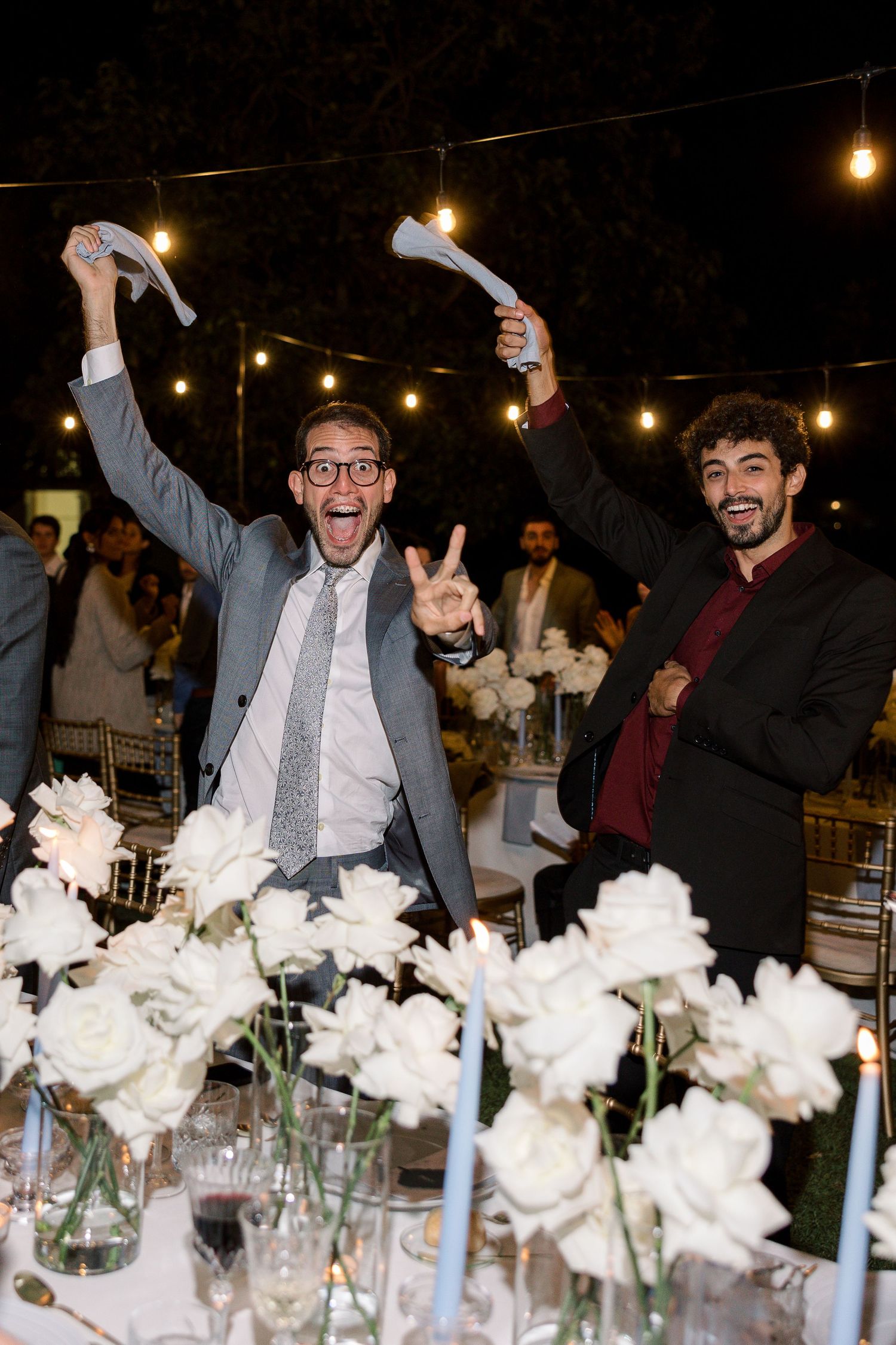 Wedding reception guests celebrate joyfully among white floral centerpieces under string lights.