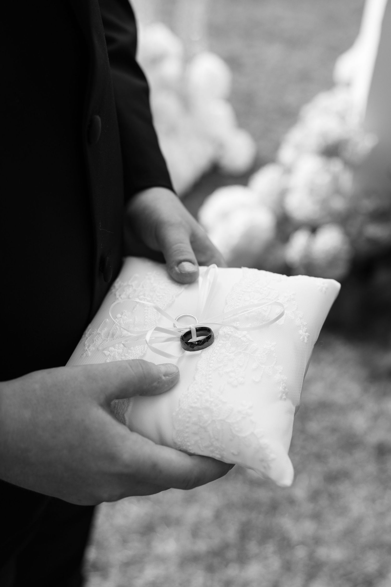 Wedding rings resting on a white lace pillow held by hands in formal attire.