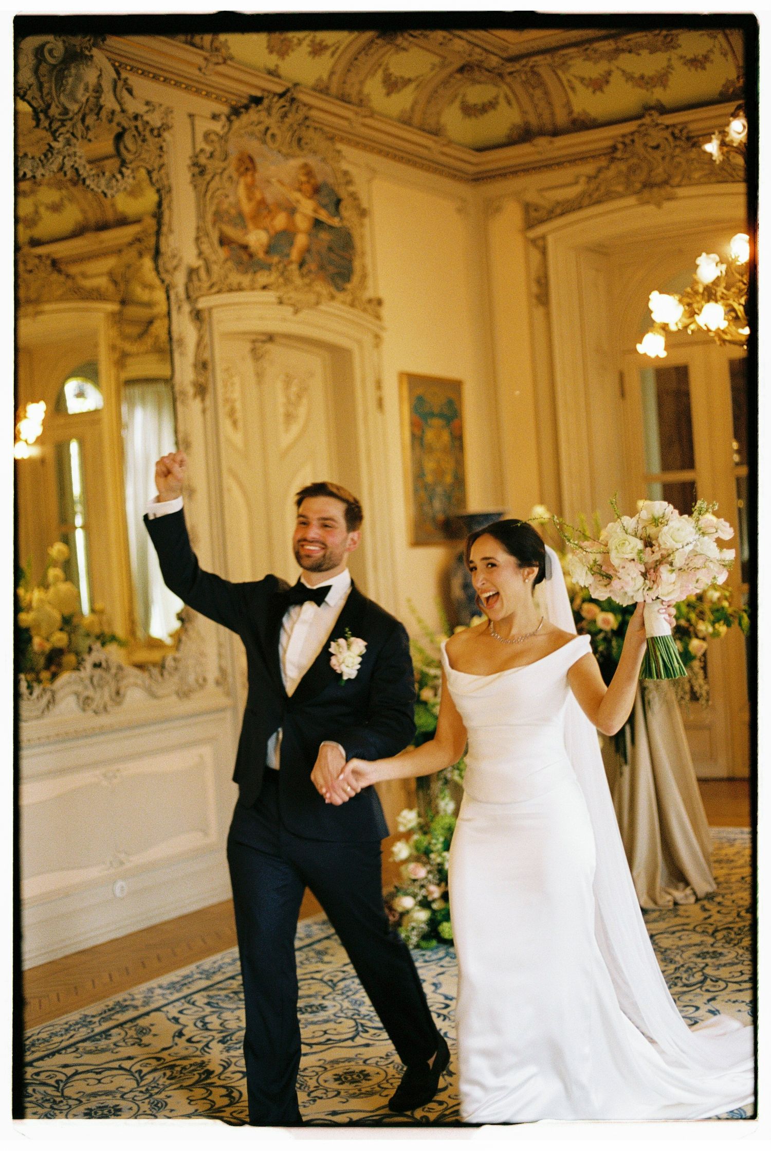 Joyful newlyweds celebrate in ornate ballroom, bride in white gown holding bouquet, groom in black tuxedo raising fist.