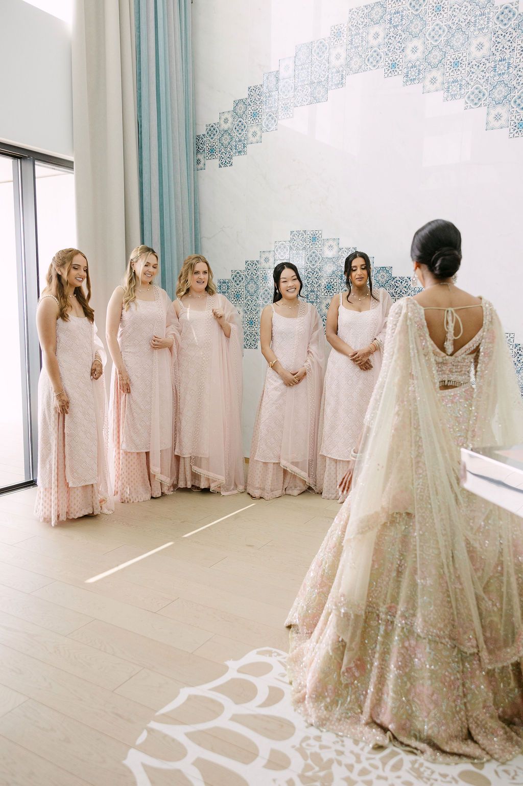 Bridesmaids in blush pink dresses stand together admiring a white wedding gown in a bright room with blue wallpaper.