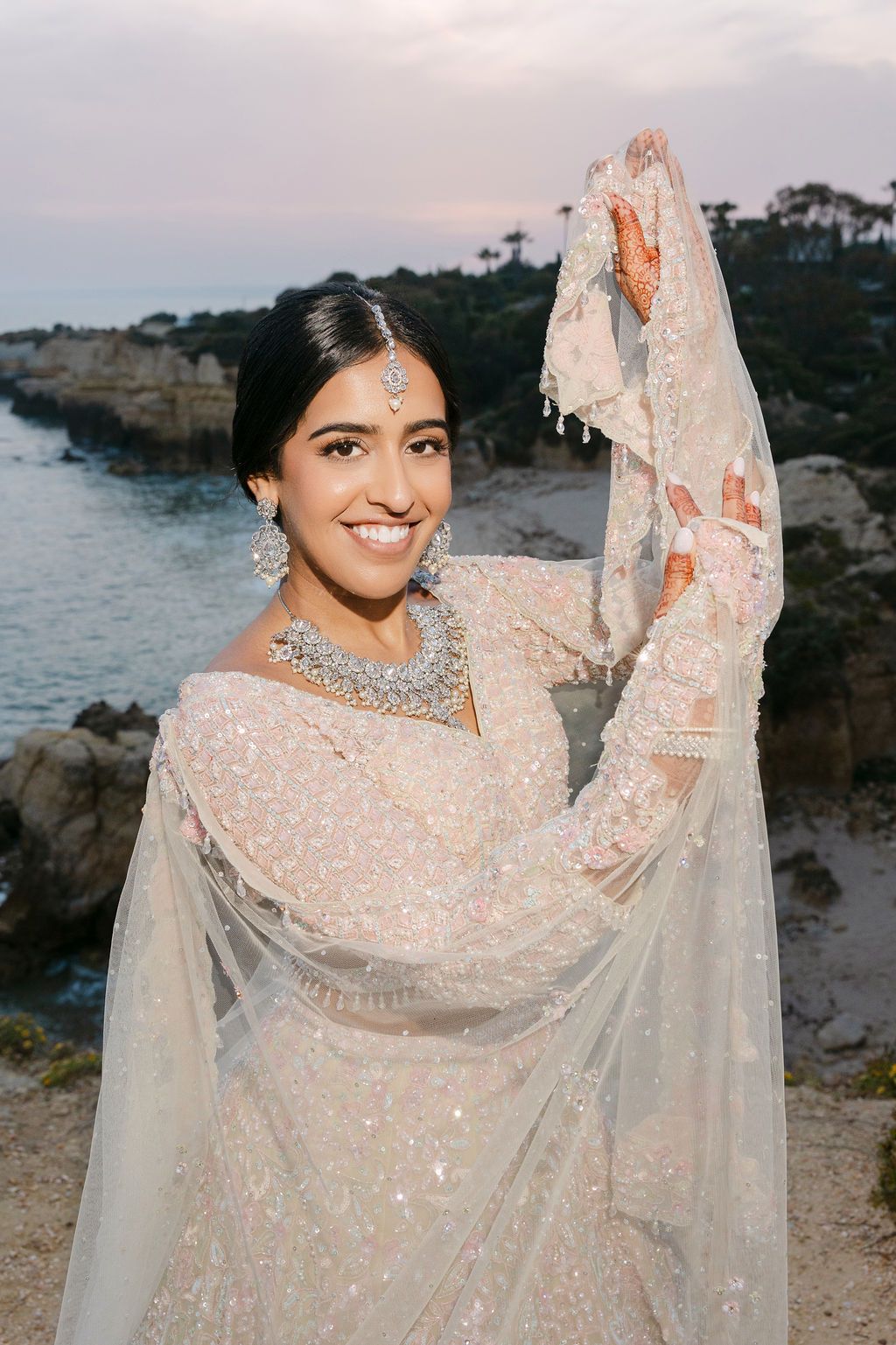 Bride in sparkling white lehenga and jewelry poses against coastal sunset backdrop.