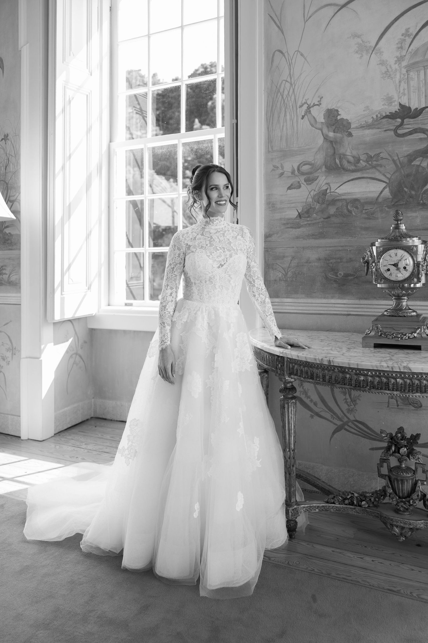 Elegant bride in long-sleeved lace wedding gown poses by window in room with decorative wall murals.