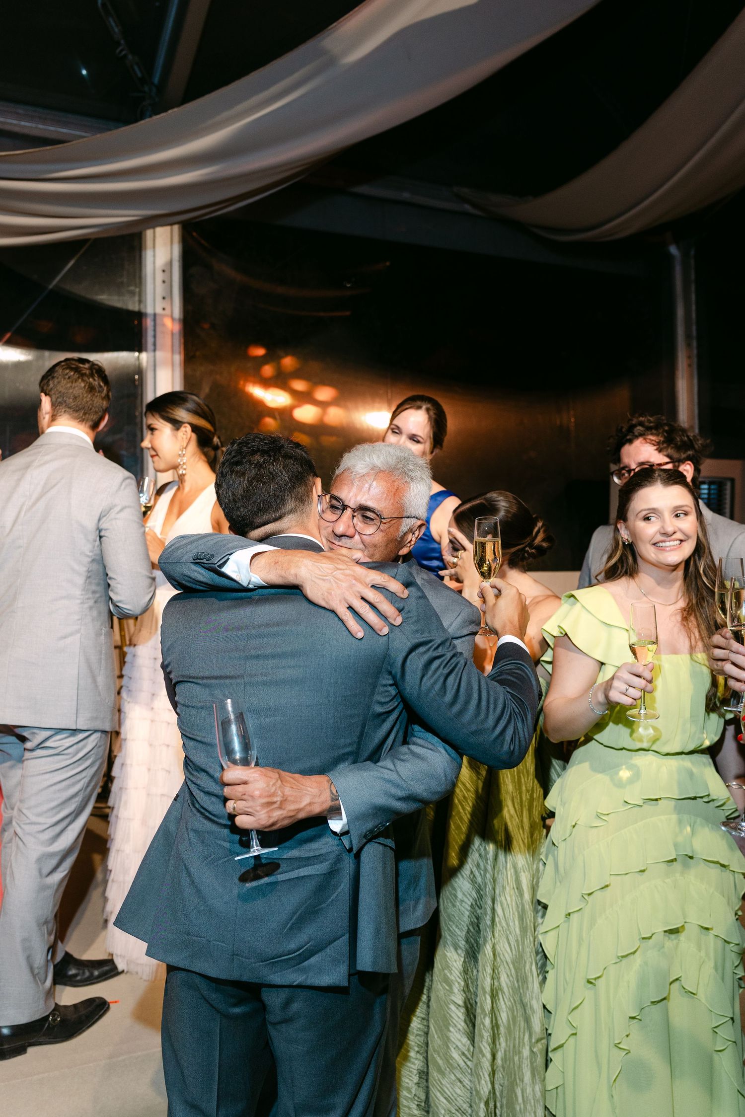Wedding guests in blue suits and mint green dresses embrace on the dance floor during a celebration.