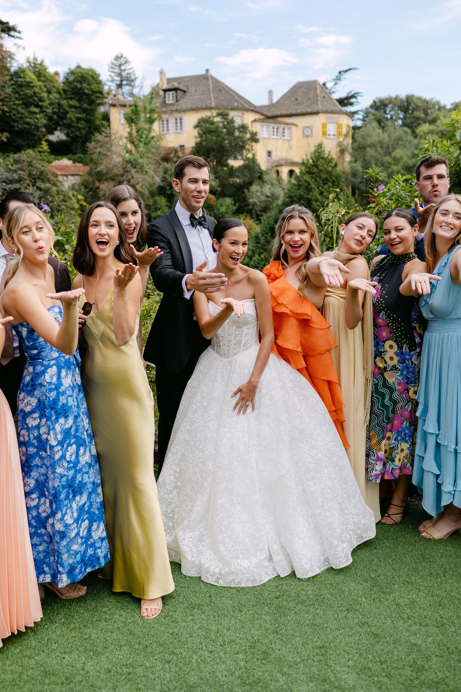Wedding party strikes fun poses on green lawn in colorful attire.