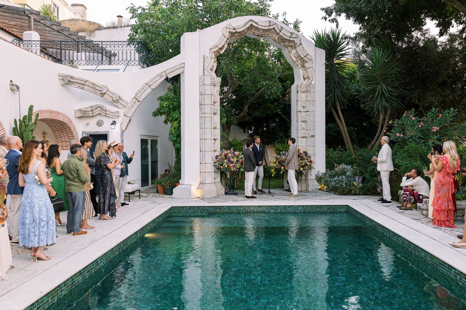 Elegant outdoor wedding ceremony beside a pool with ornate white arch, lush greenery, and guests in a Mediterranean garden setting.