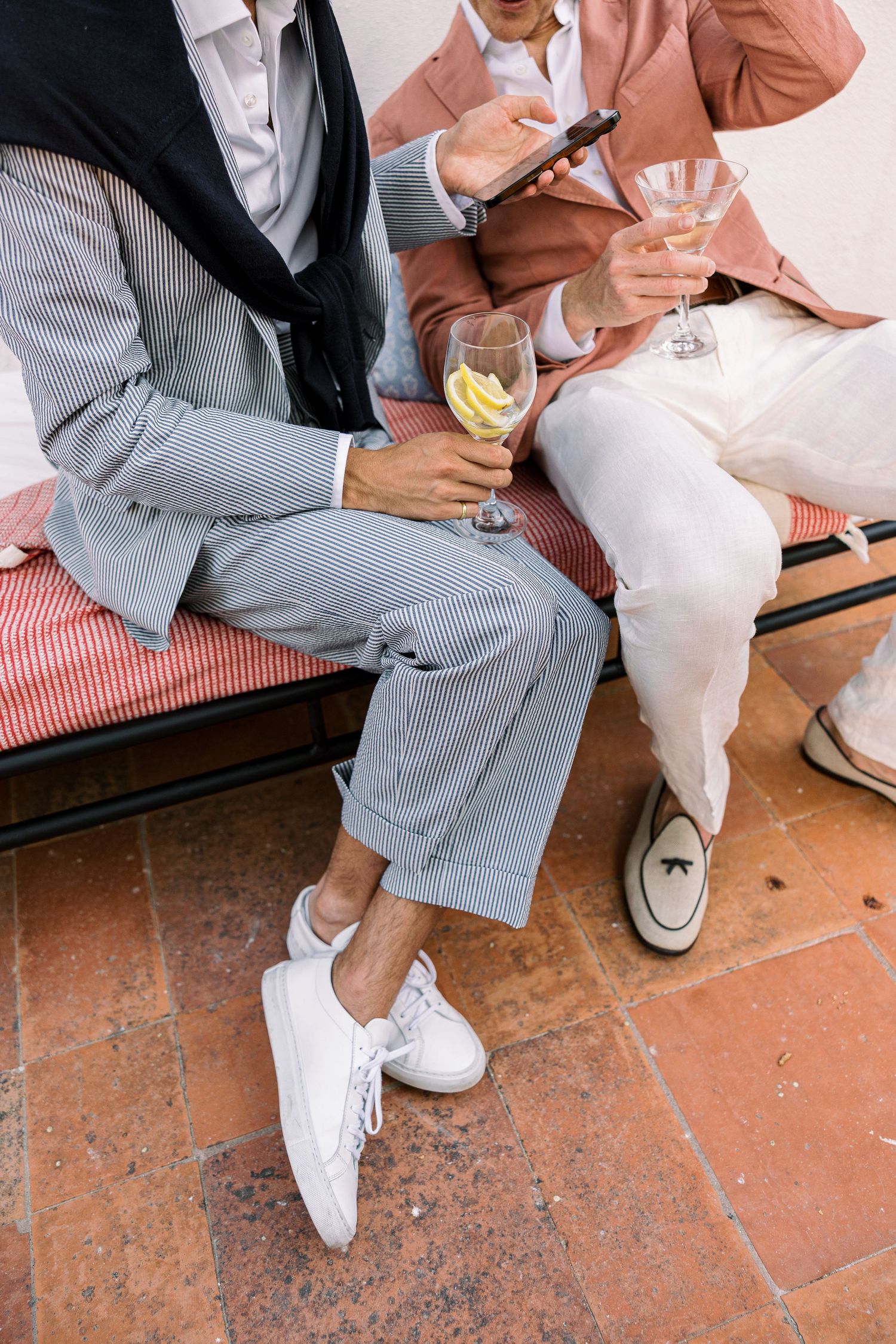 Two men in elegant suits sit on a bench holding wine glasses at an upscale outdoor event.