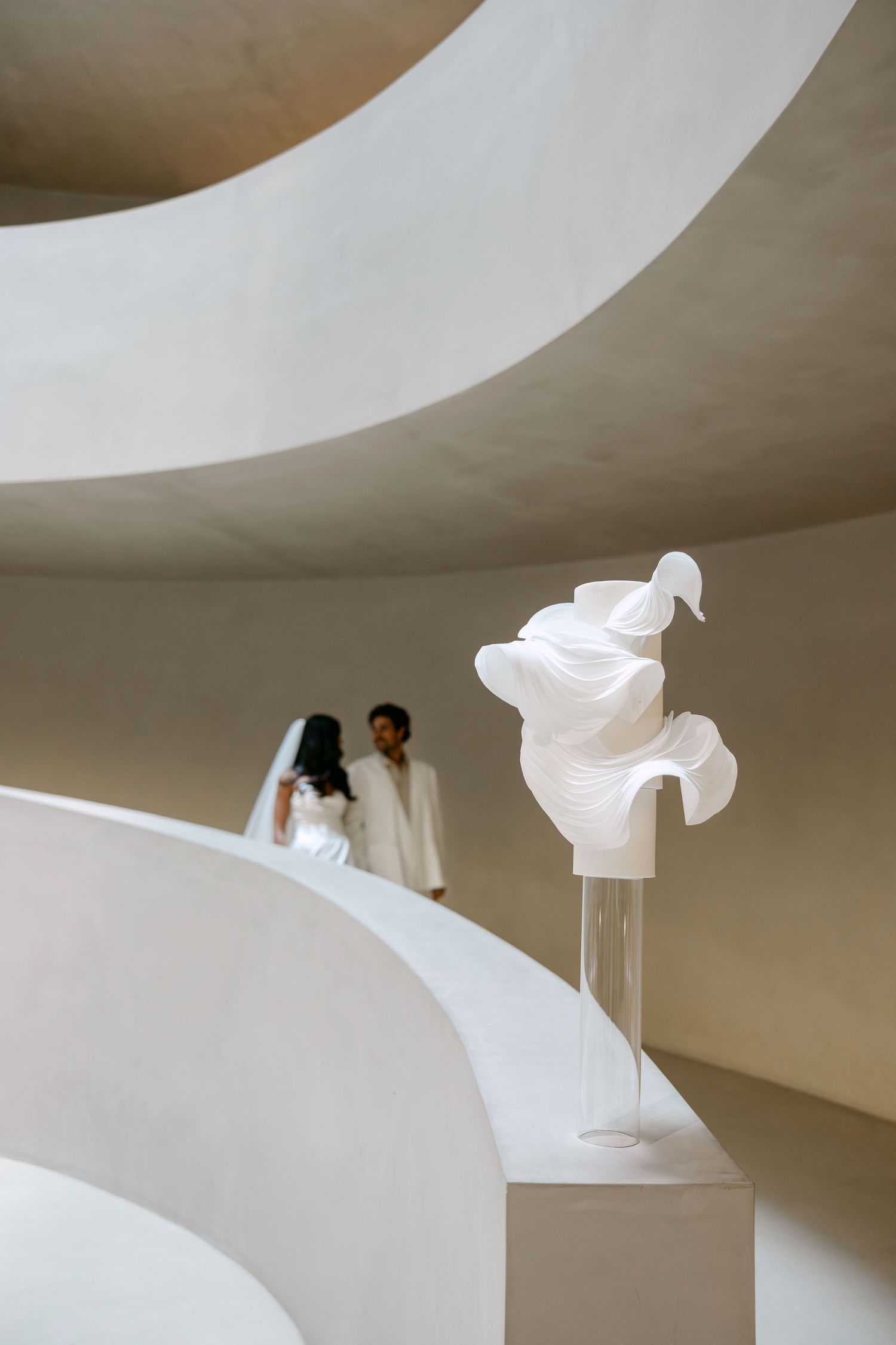 A bride and groom stand in a curved white architectural space beside an elegant flowing white fabric sculpture on a pedestal.