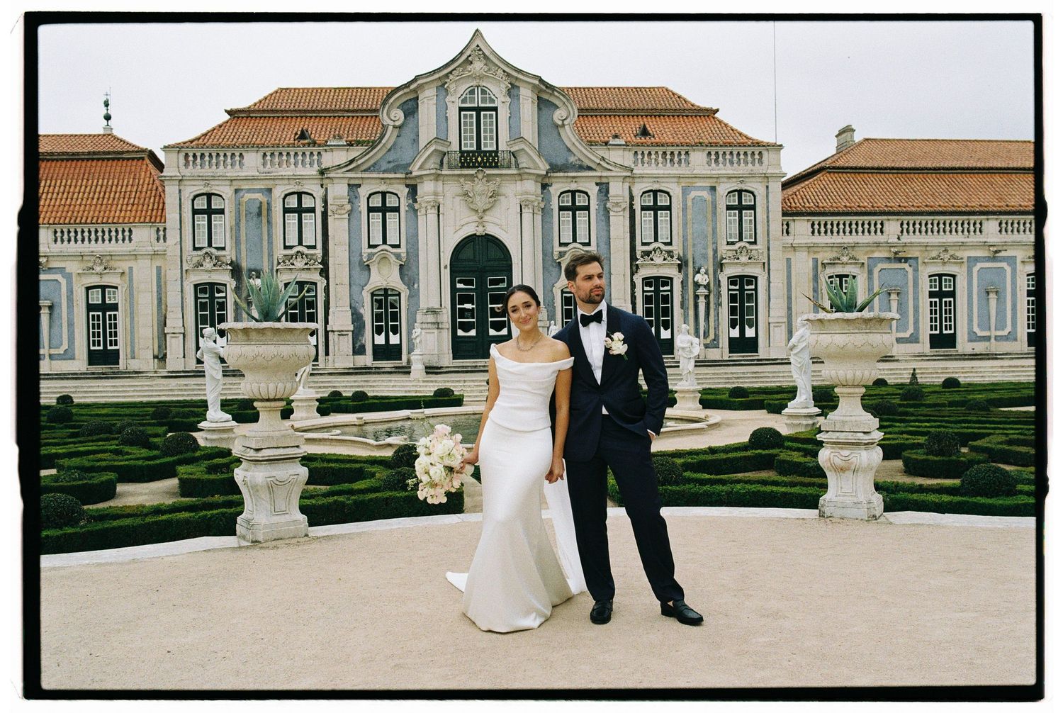 Bride and groom pose before an elegant baroque palace with manicured hedges and stone urns at their wedding.