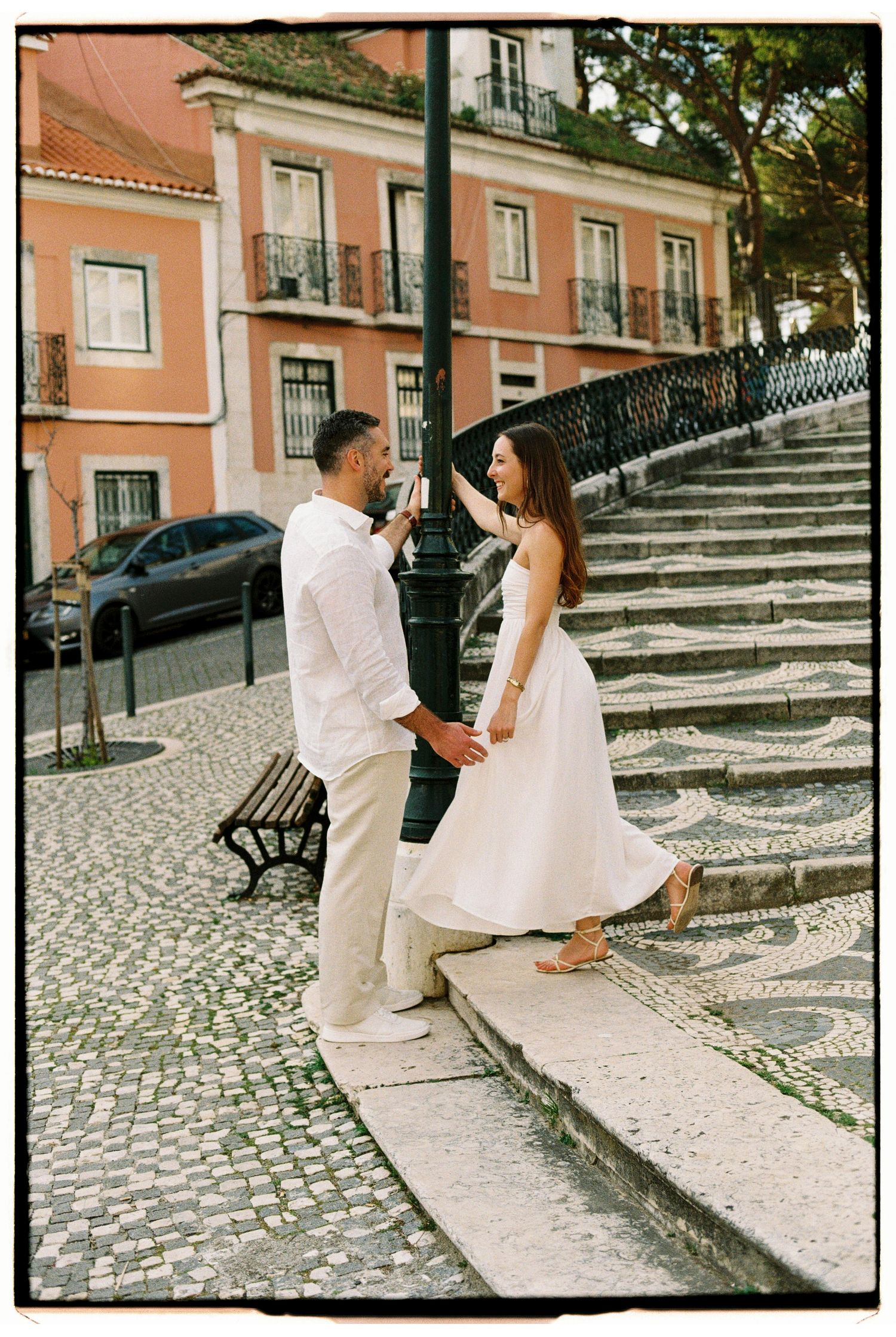 Couple holding hands on cobblestone steps in Lisbon, woman in white dress, man in white outfit, pink building backdrop.