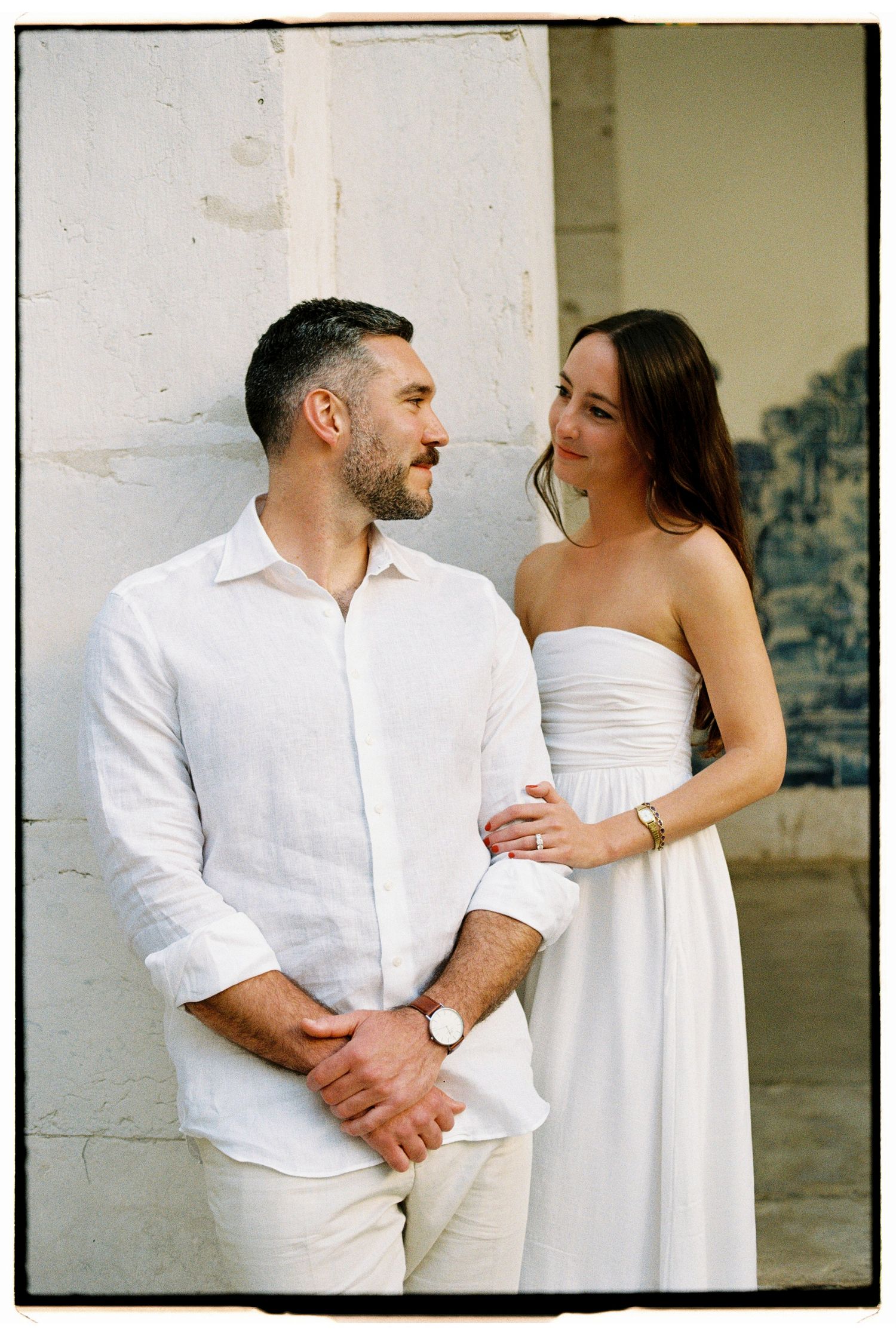 Couple in matching white outfits posing by a stone wall, woman in strapless dress touching mans arm.