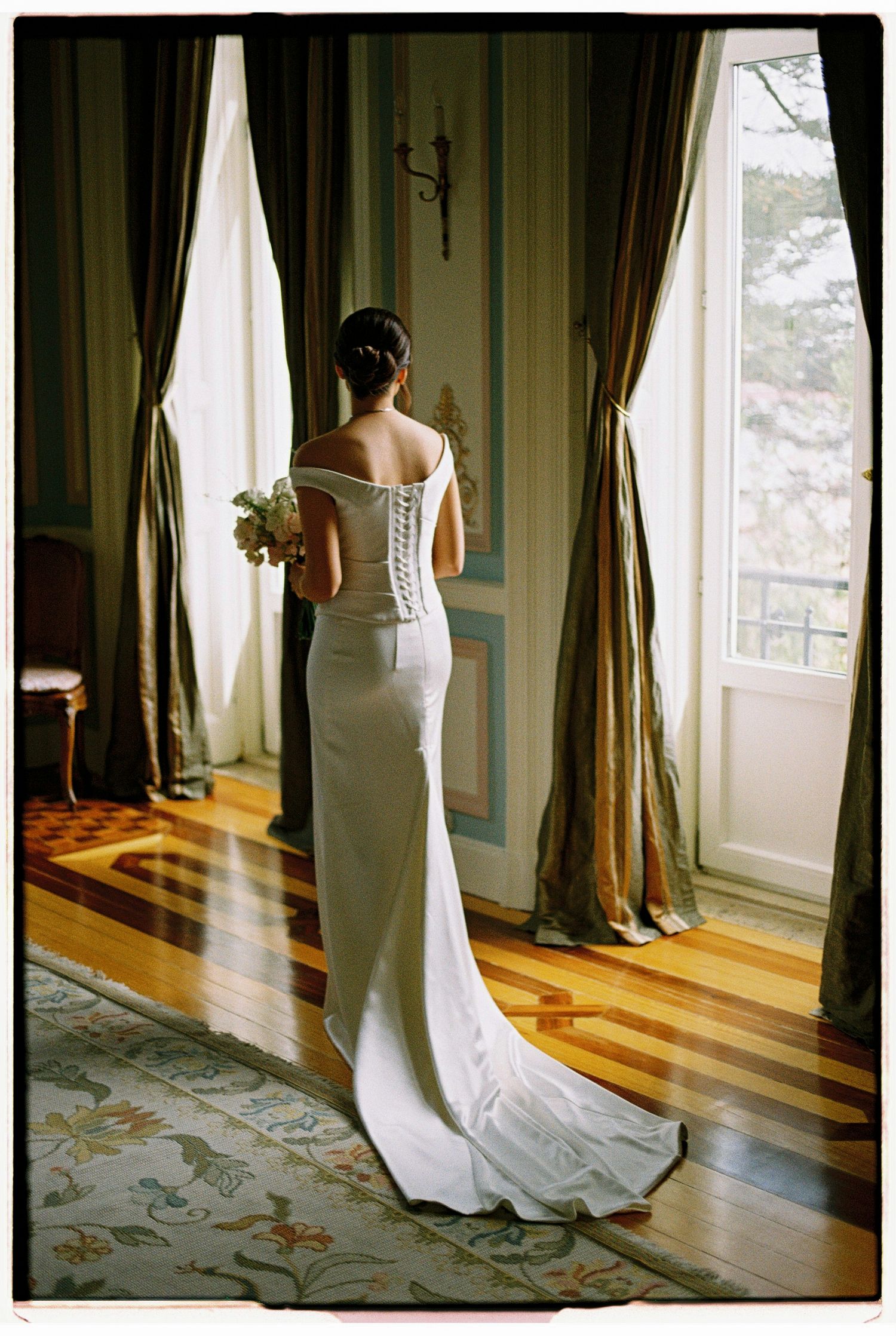 Bride in white off-shoulder gown with corset back and train stands by tall window with gold drapes.