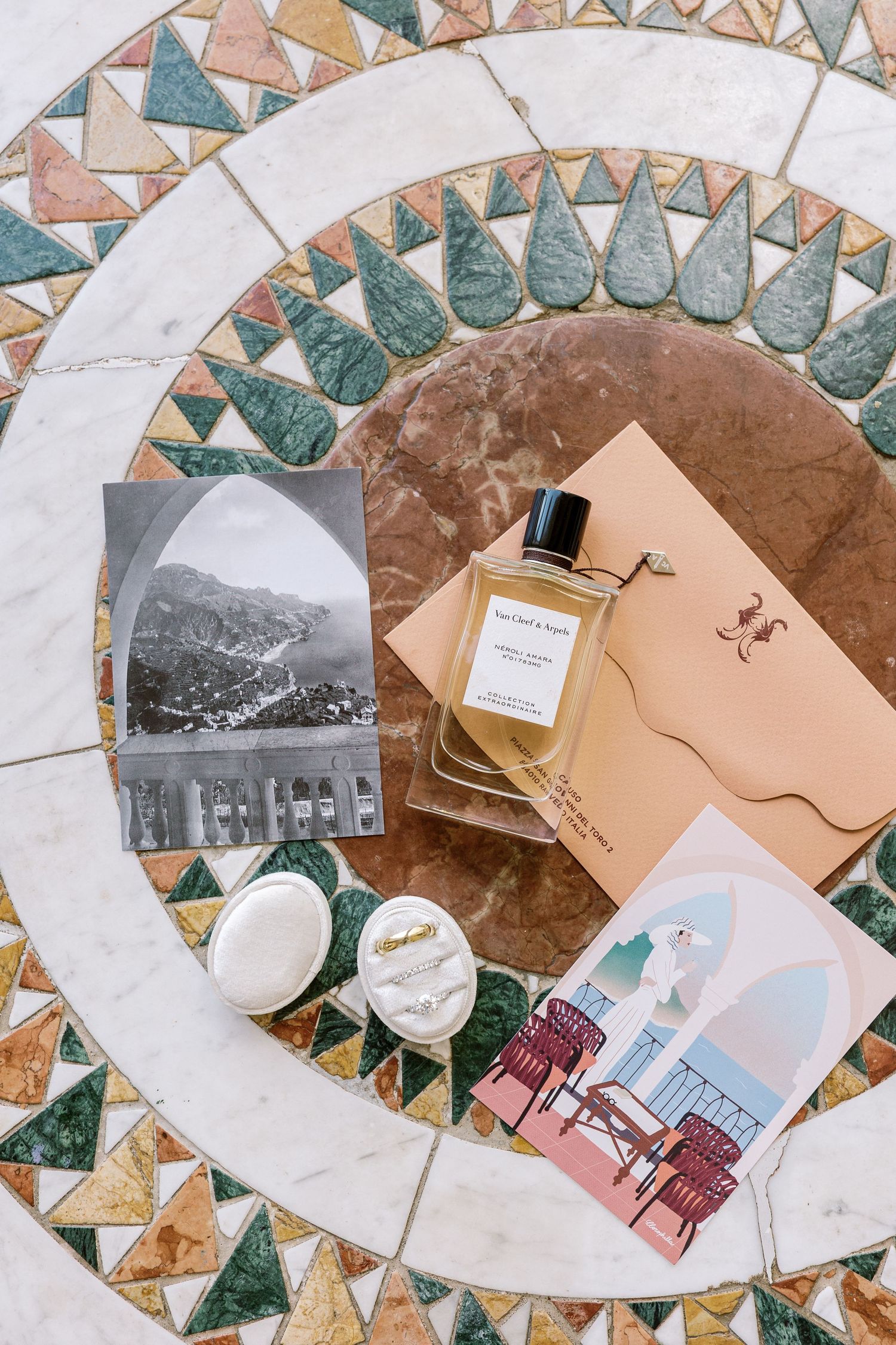 Flat lay of perfume bottle, postcards, and cream jars on ornate mosaic marble table.