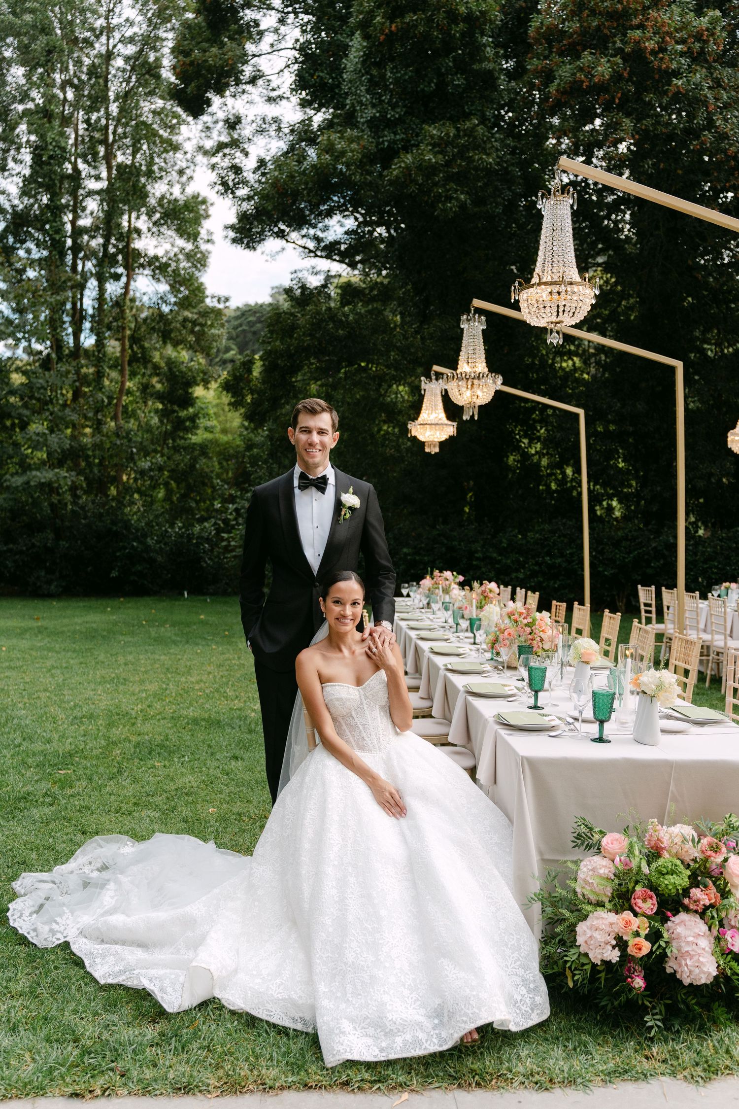 Newlyweds pose by elegant outdoor reception table with crystal chandeliers and floral arrangements.