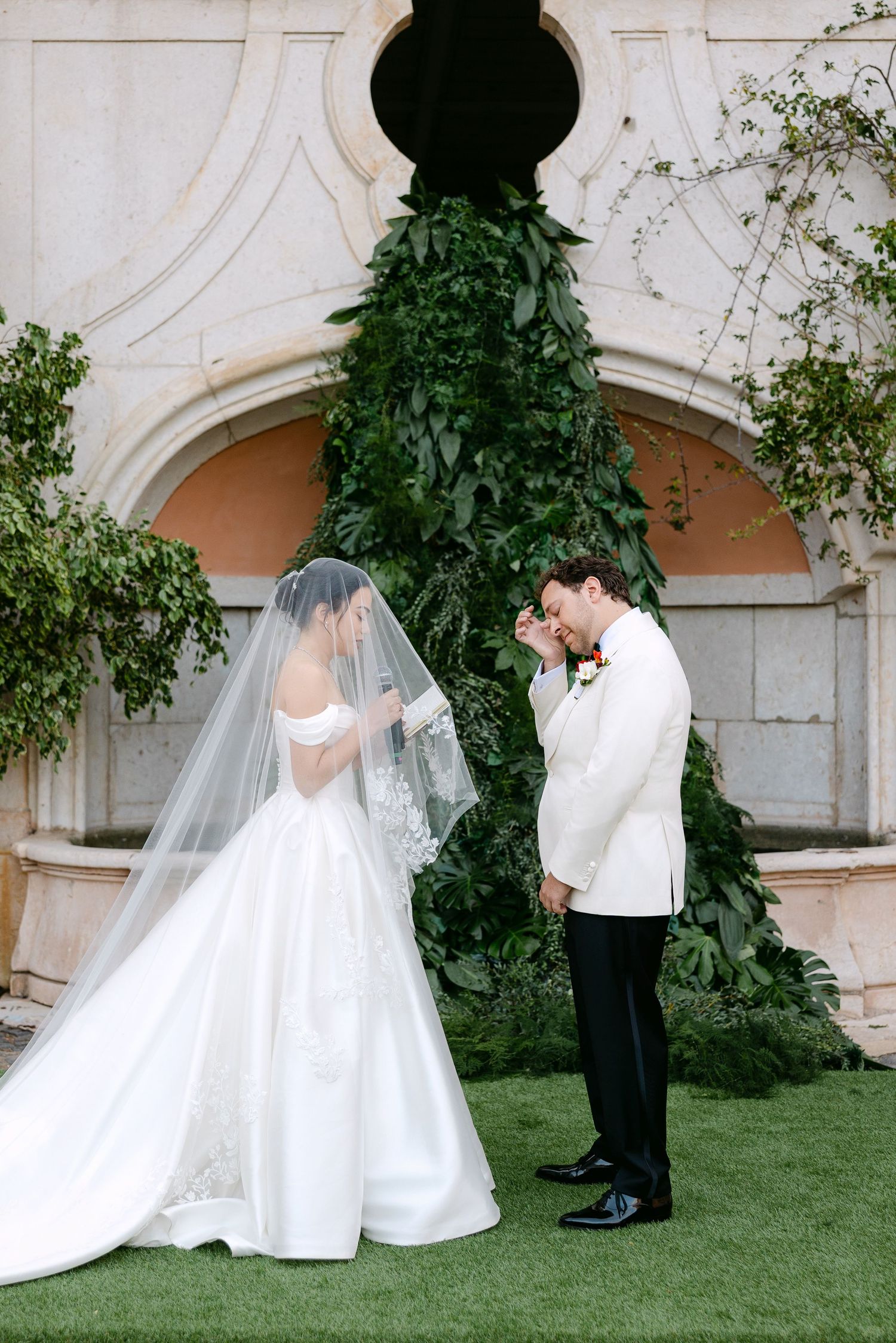A romantic wedding couple shares intimate moments in a garden setting with elegant architecture in the background.