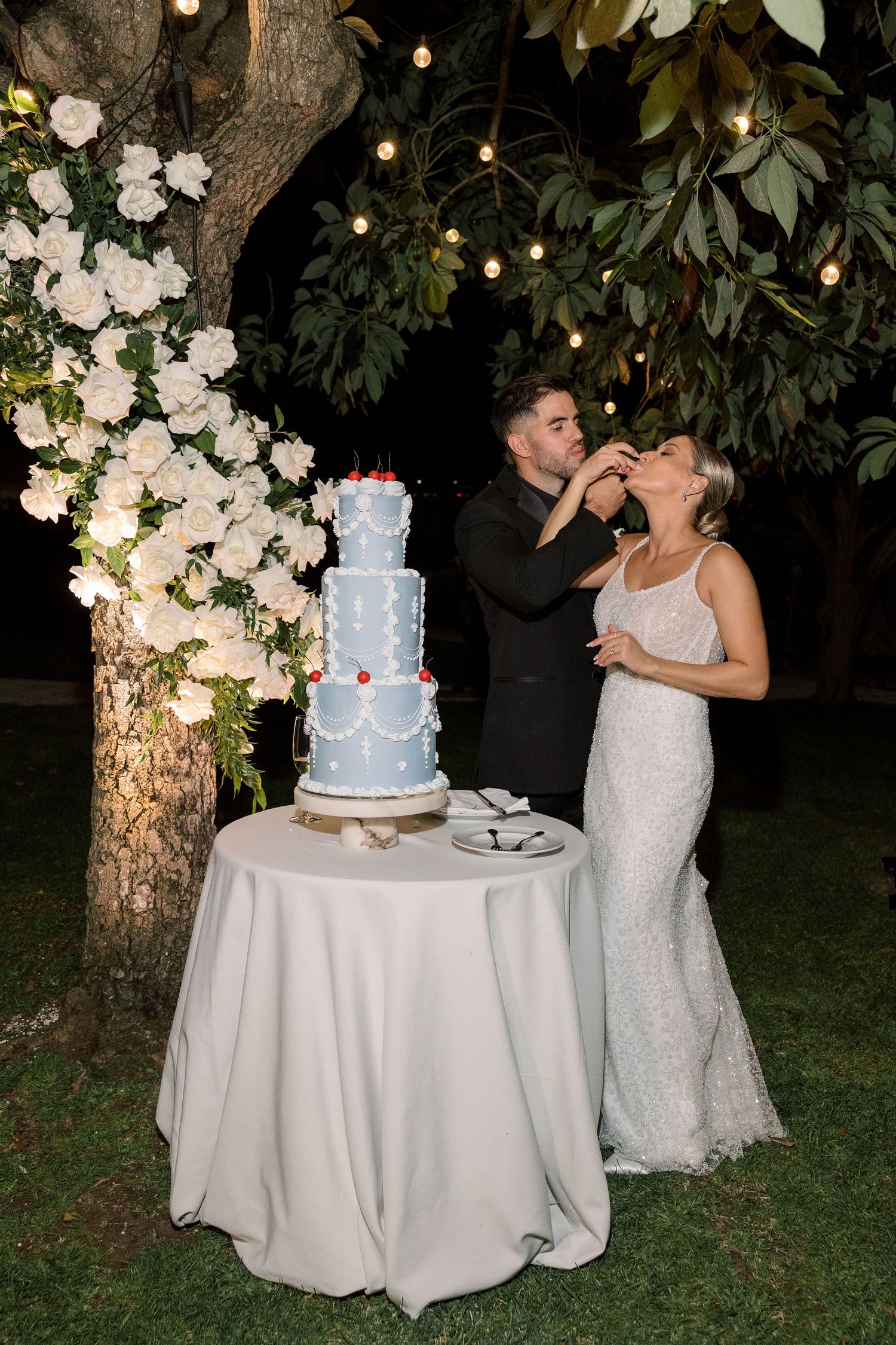Wedding cake display with white roses and string lights in an outdoor garden setting.