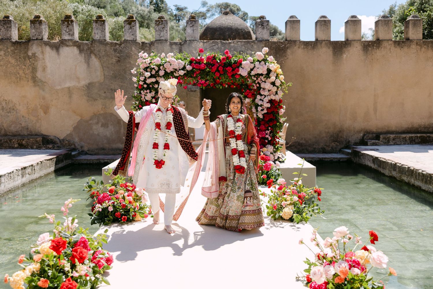 Ornate floral wedding arch with red and white flowers set against stone walls beside a reflecting pool.