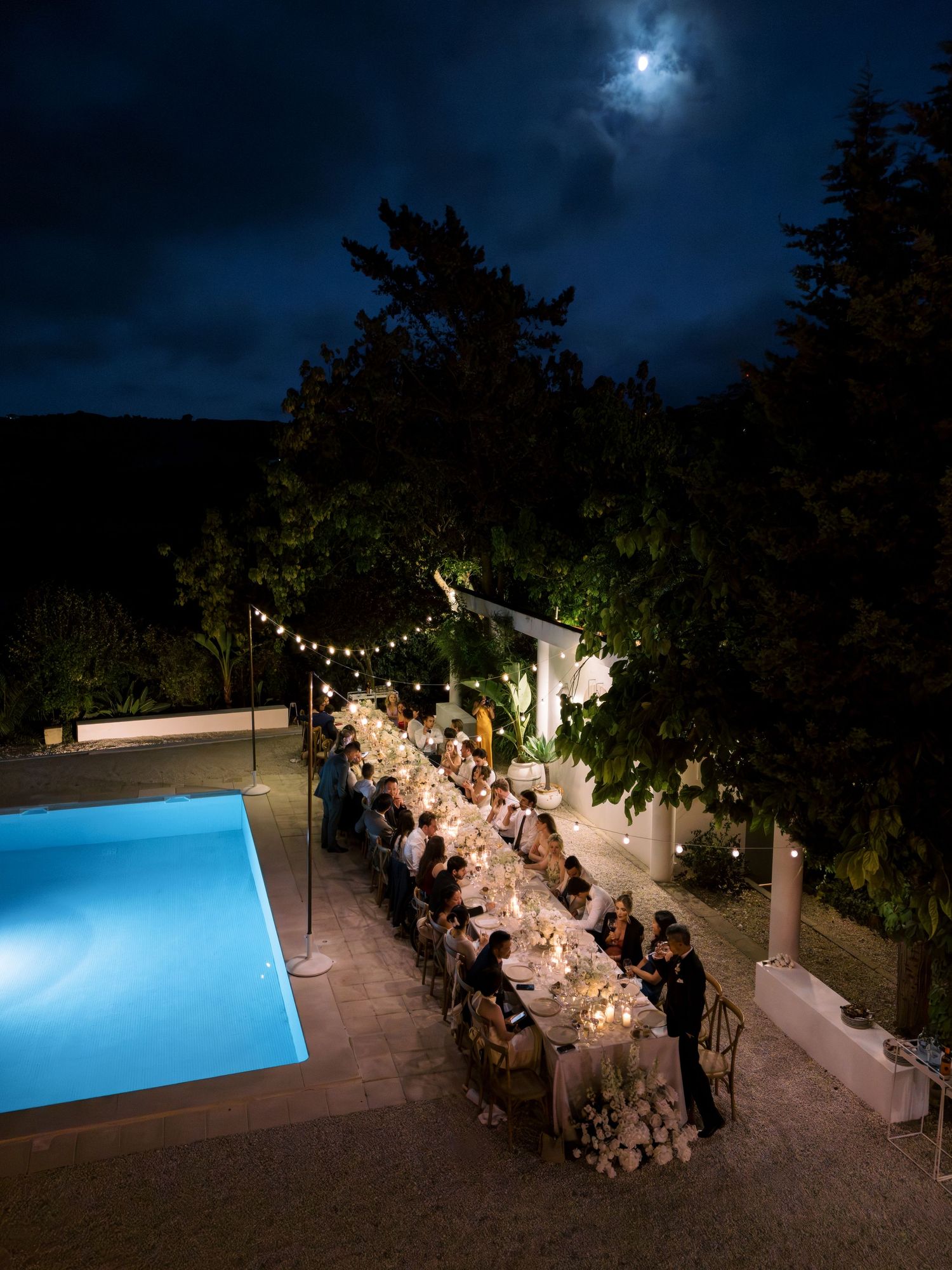 Elegant outdoor dinner party by a pool at night, with string lights and moonlight illuminating a long decorated table.