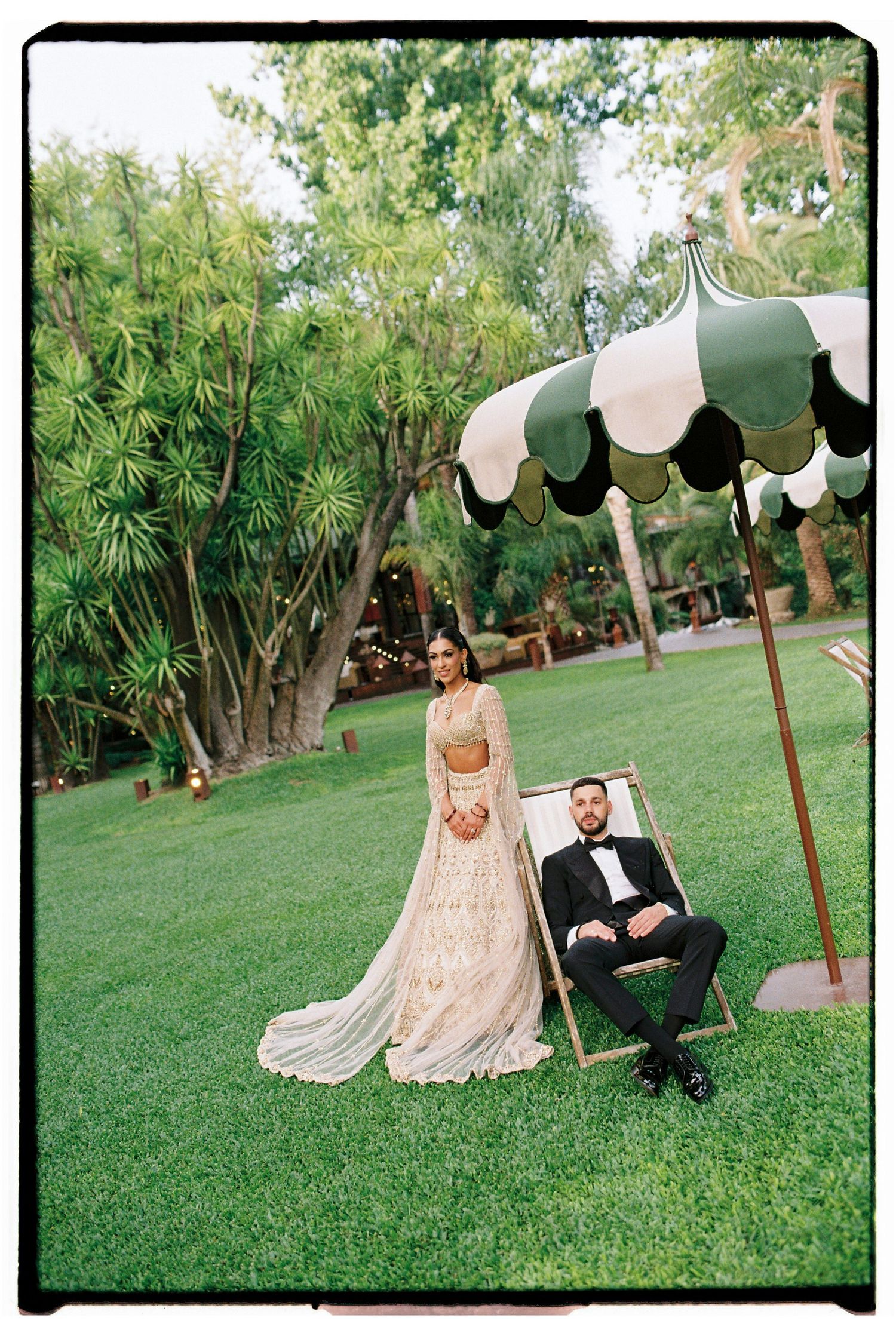 Bride in gold lehenga and groom in tuxedo pose under striped green umbrella on lush lawn at outdoor wedding.