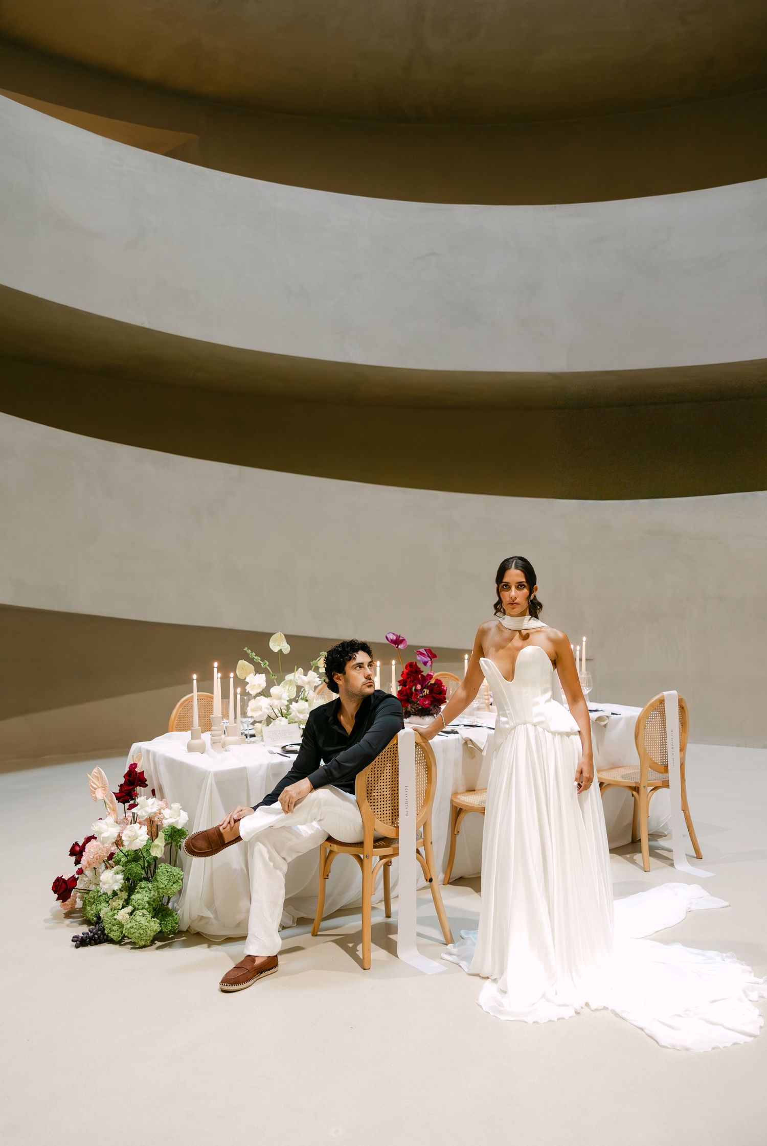 Bride and groom pose at elegant sweetheart table with candles and floral arrangements in curved venue.