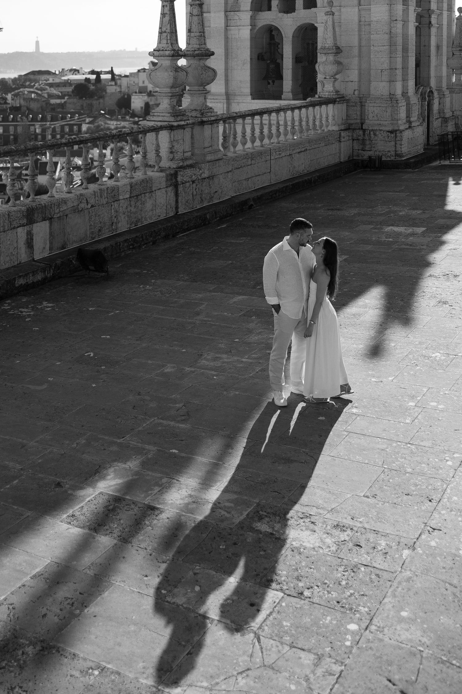 Black and white photo of a romantic couple in white attire sharing a kiss on a historic stone terrace with long shadows.