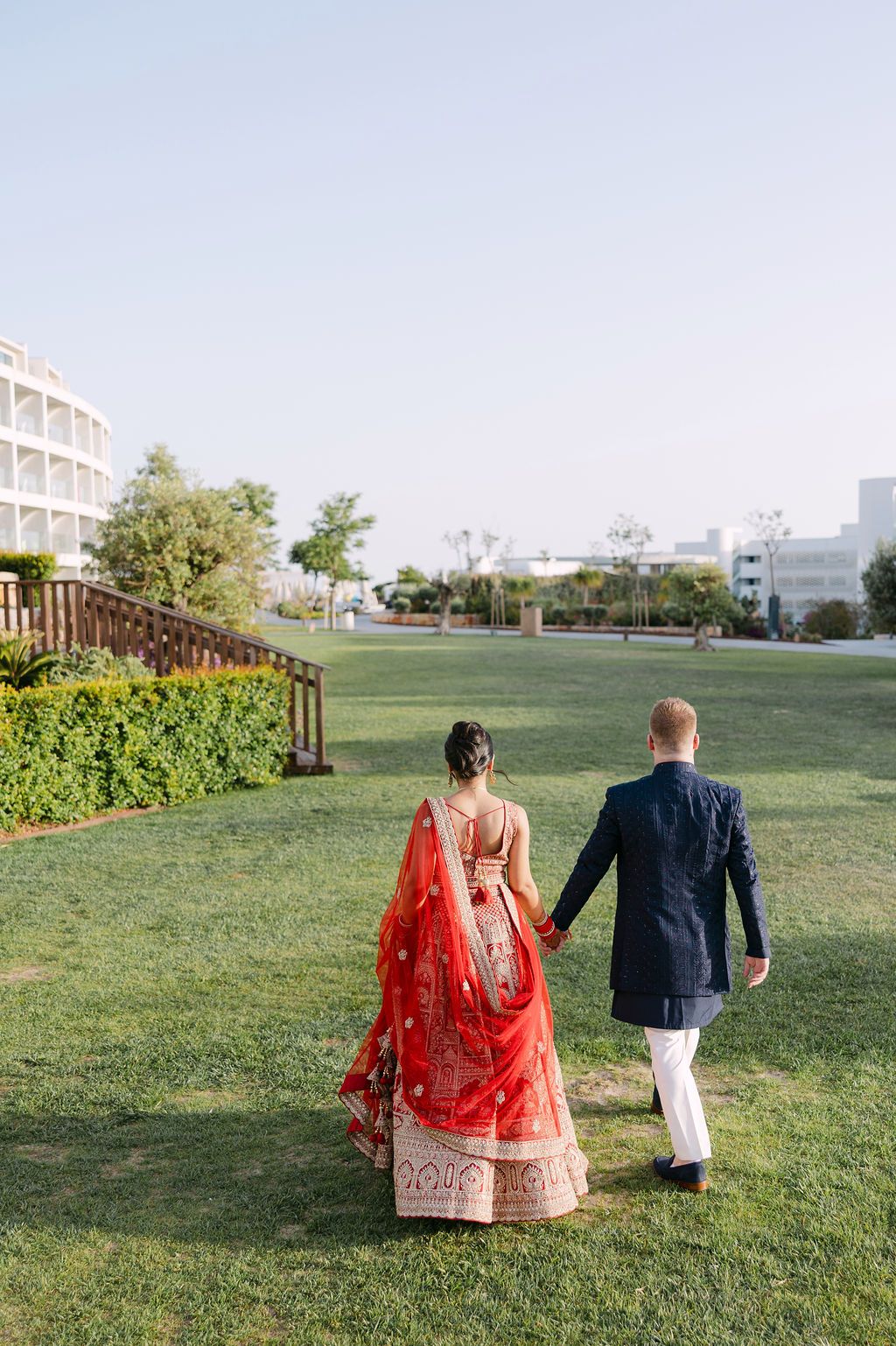 A couple walks on a grassy lawn, with the bride wearing a traditional red lehenga and groom in a navy suit.