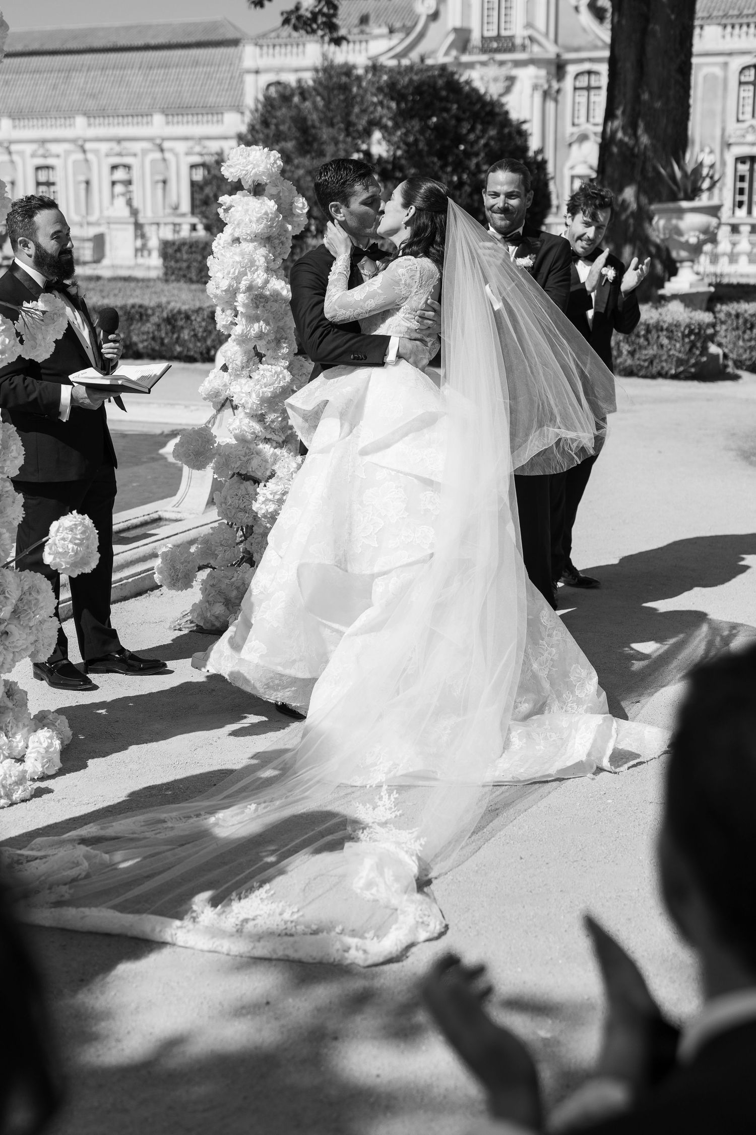 Bride and groom share first kiss surrounded by guests and floral arch in black and white wedding photo.